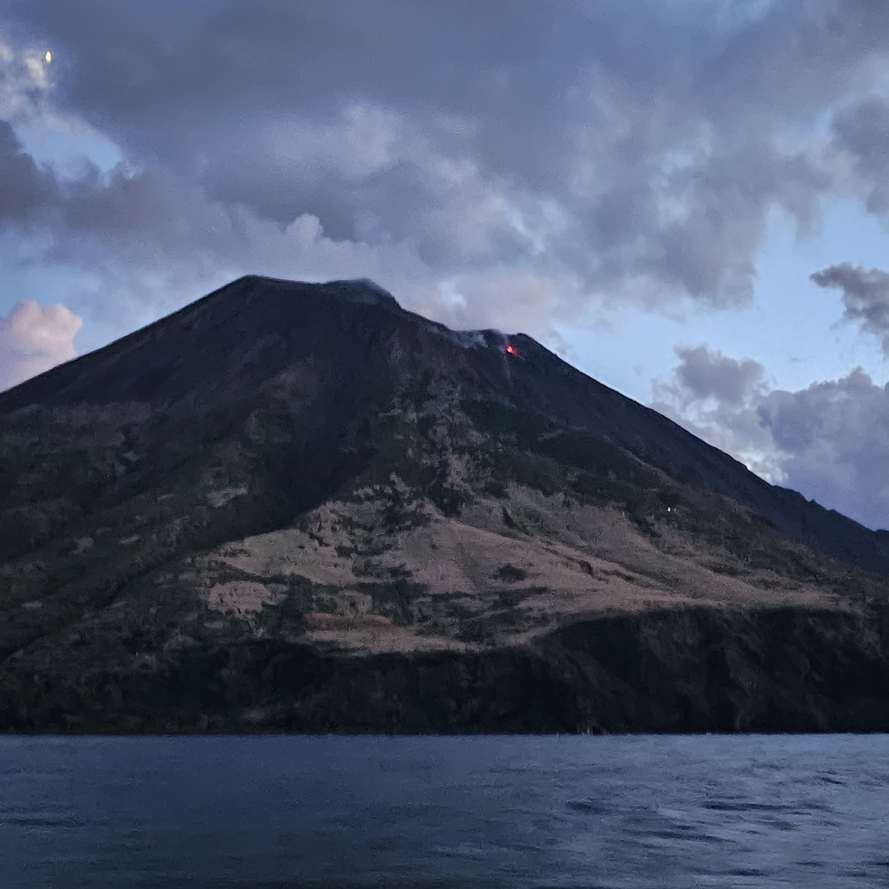 A volcanic mountain at dusk with a small red glow near the summit above the sea.