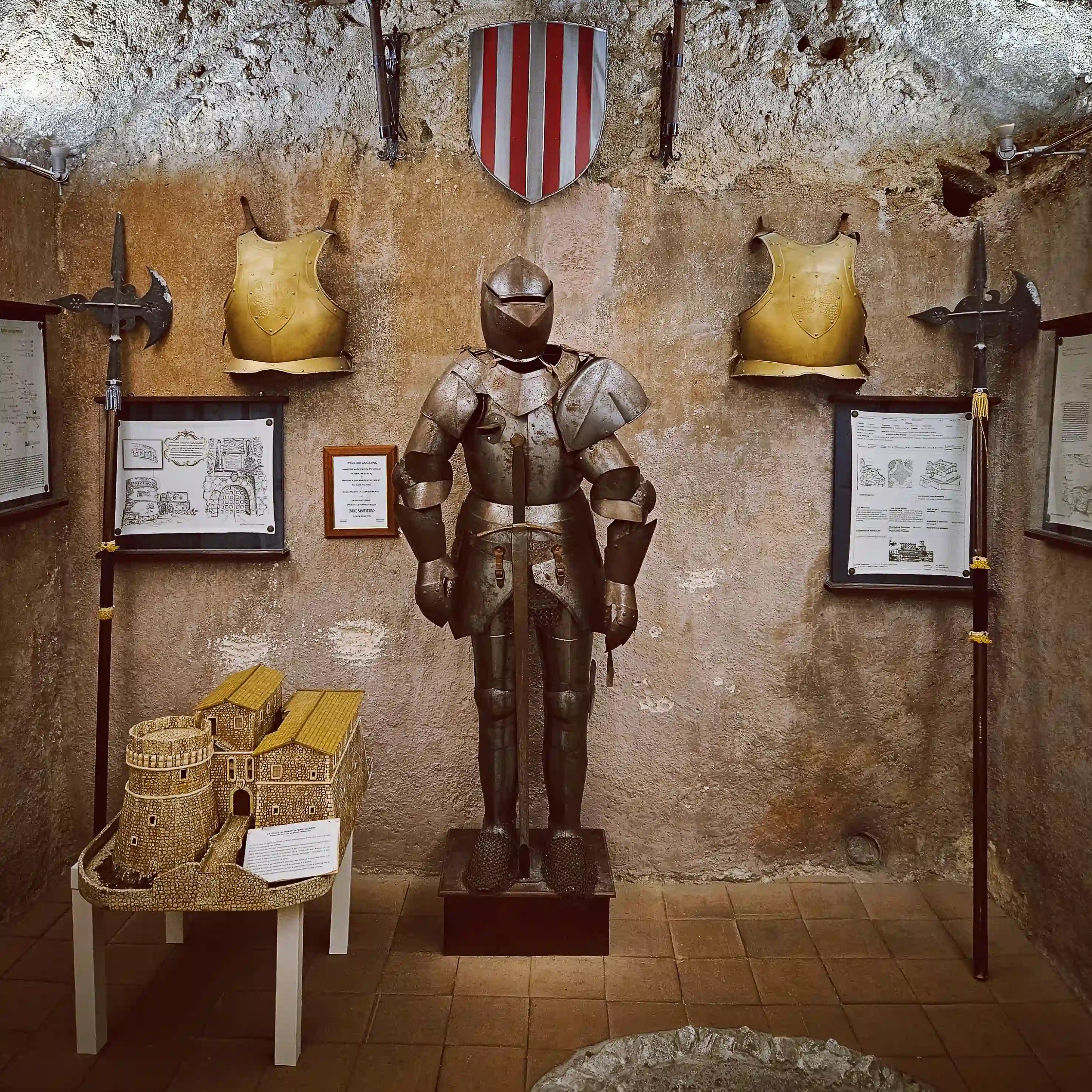 A suit of armor stands in the center of a room surrounded by shields, weapons, and informational displays on stone walls.