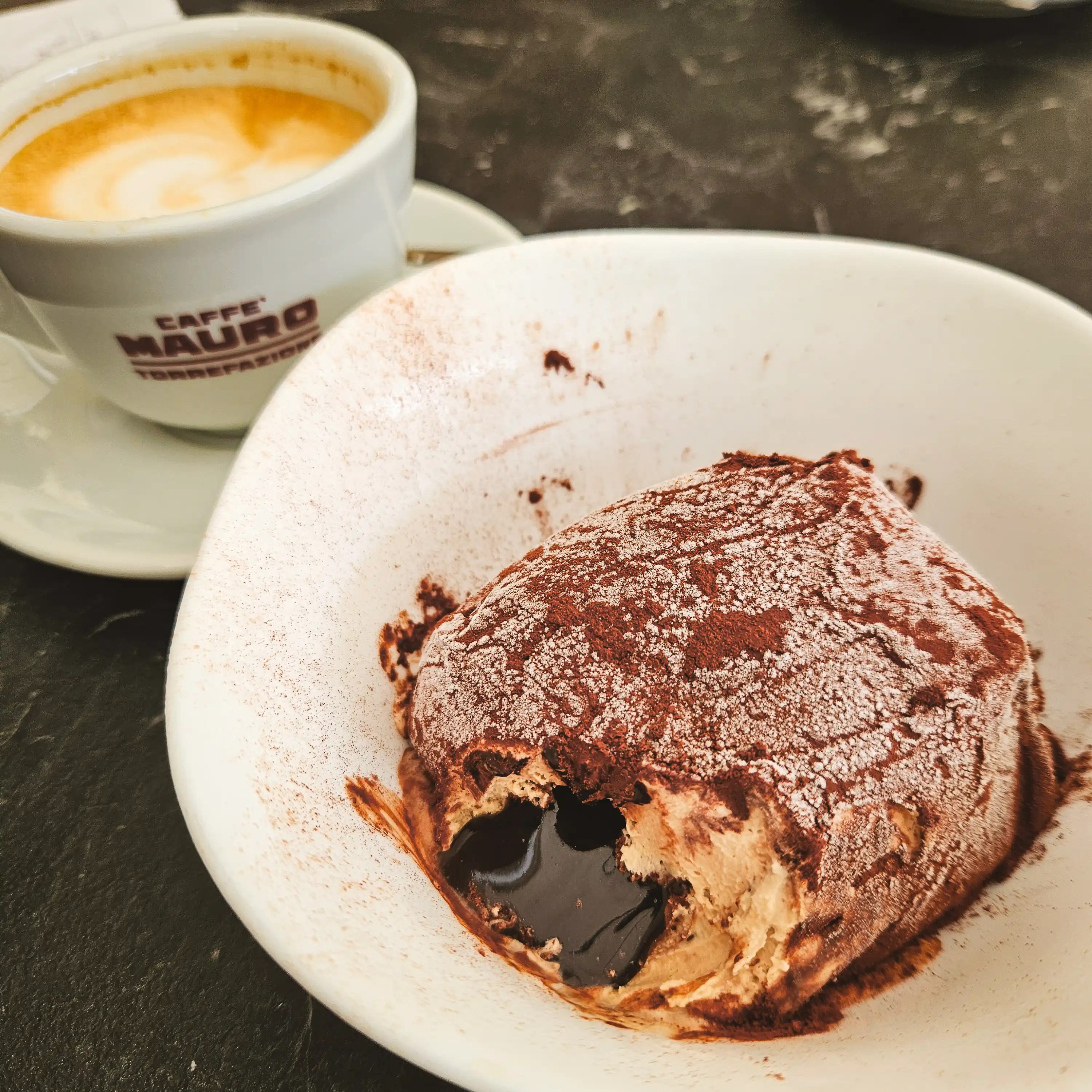 A partially cut chocolate dessert with a molten center sits on a plate beside a cappuccino.