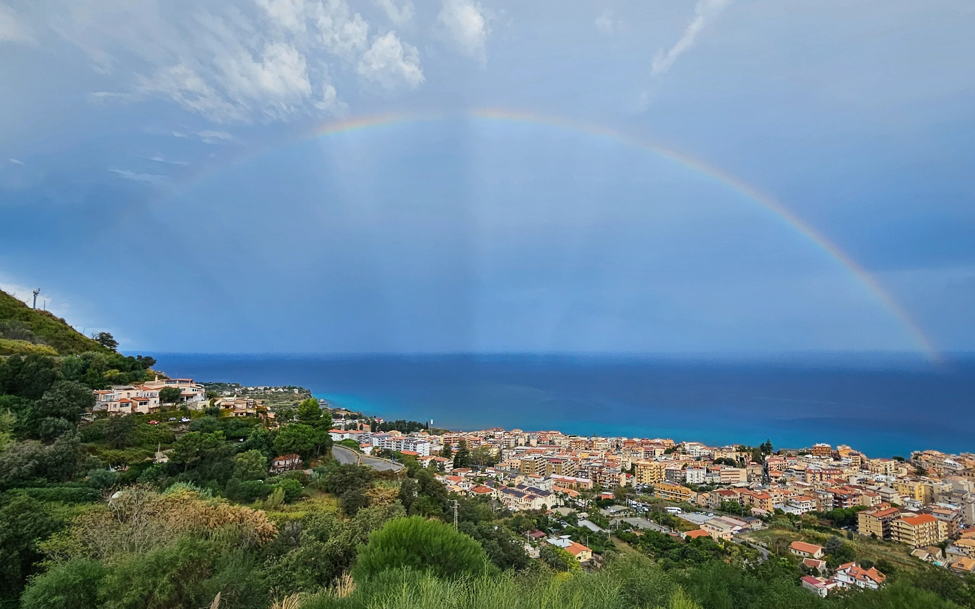 Wide view of a coastal town and sea with a rainbow arching across a cloudy sky.