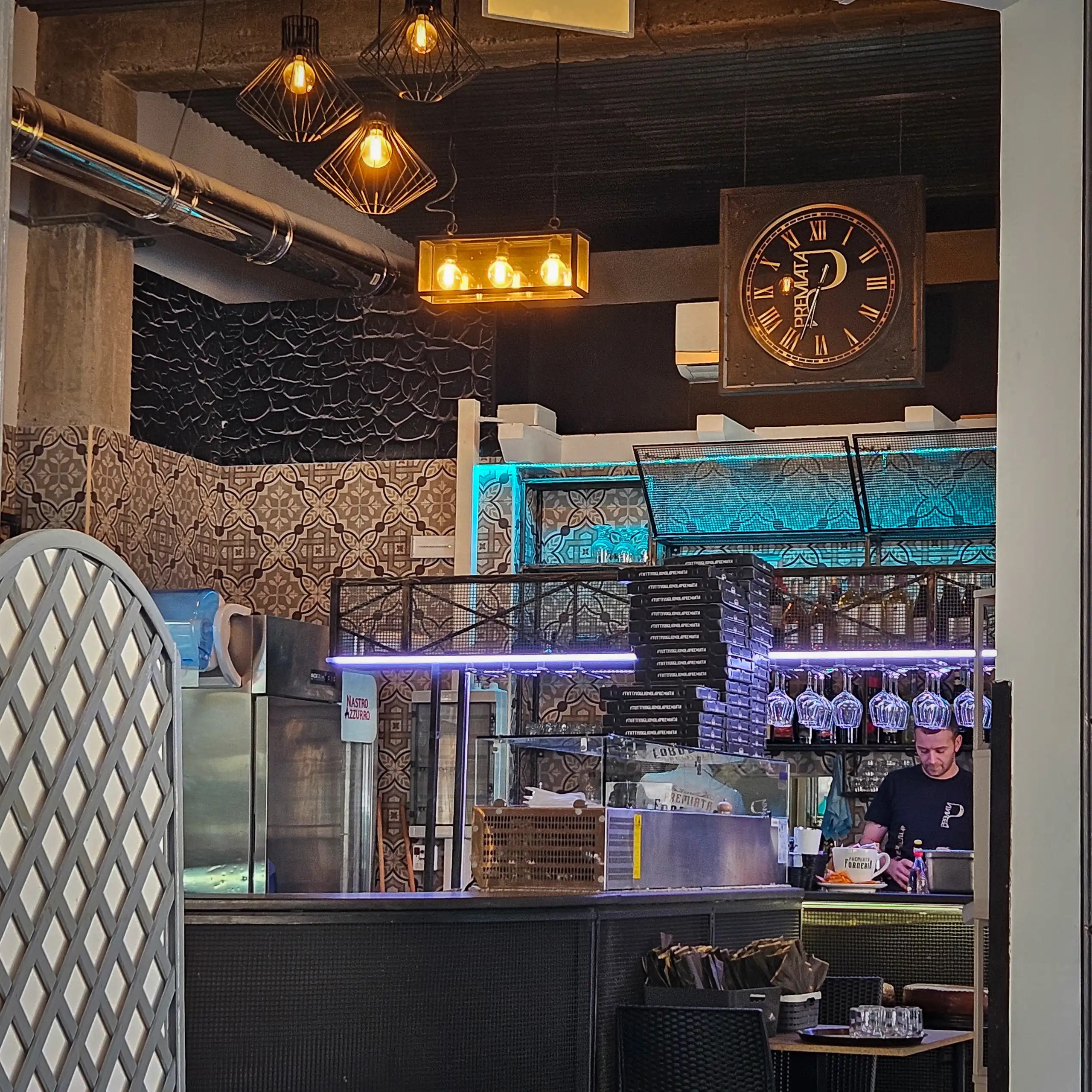 Restaurant interior with hanging lights, tiled walls, a counter with glassware, and a staff member working behind it.