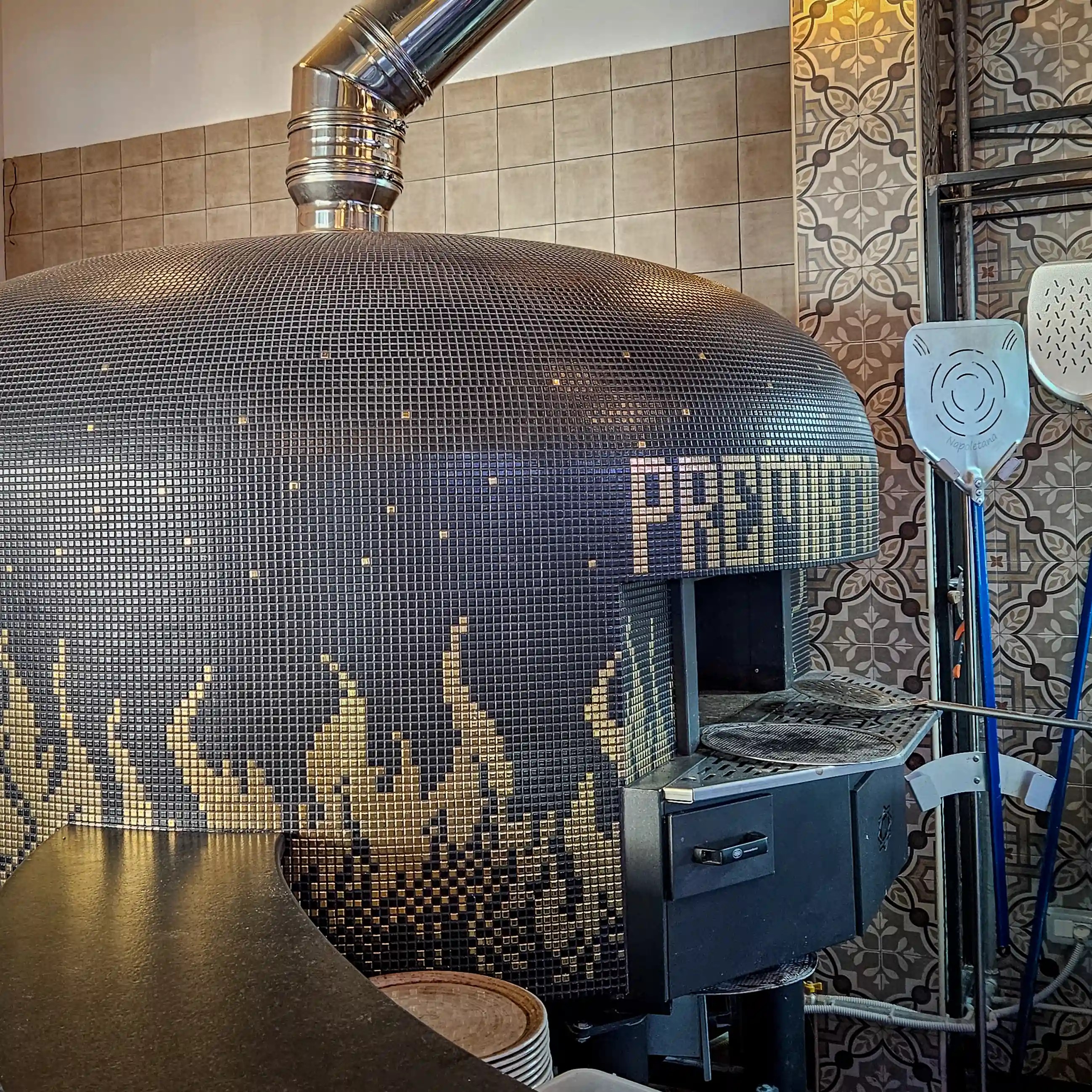 A tiled pizza oven with a metal chimney and a door opening, with pizza tools standing nearby.