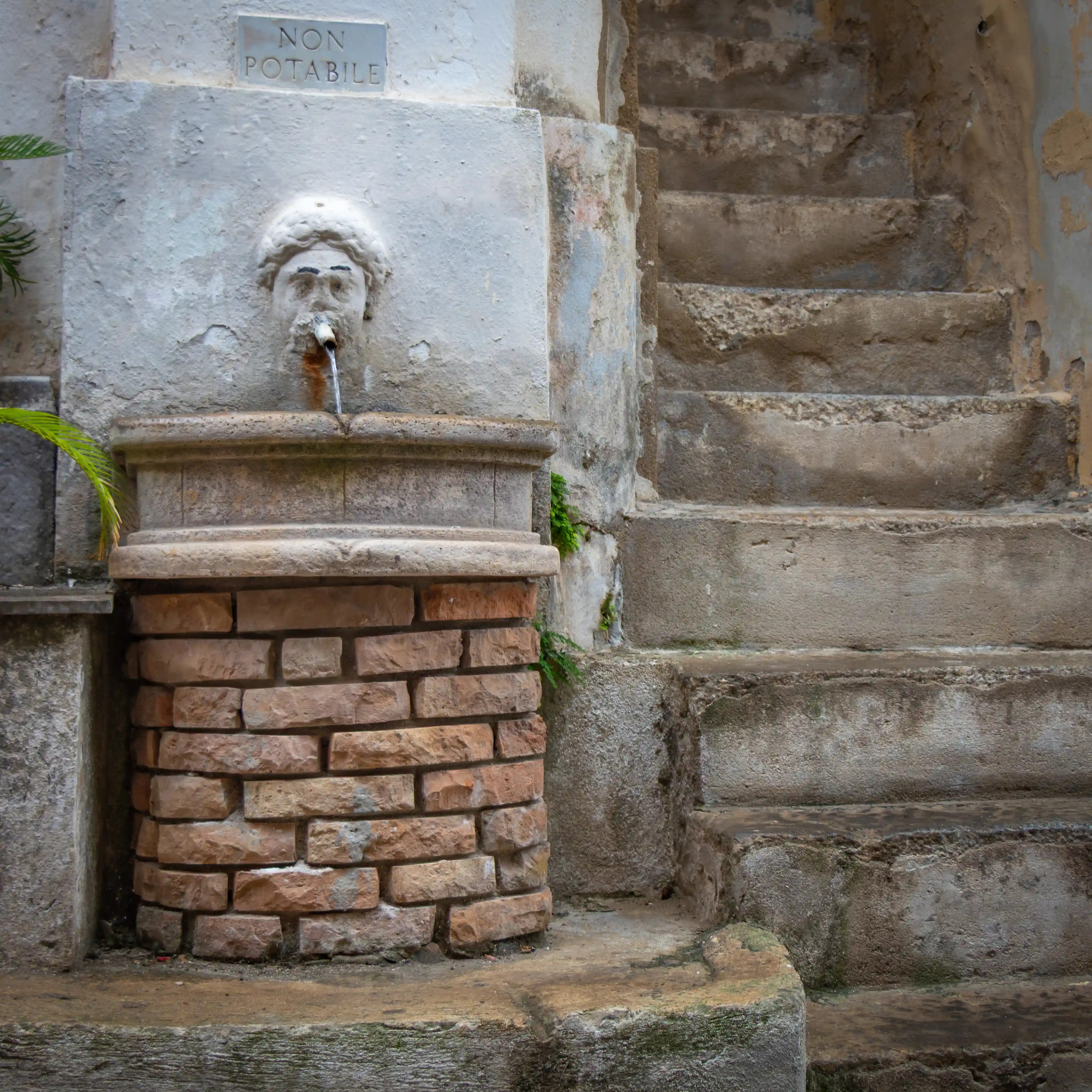 A small stone fountain with a carved face spout and a “NON POTABILE” sign above it beside worn stone steps.