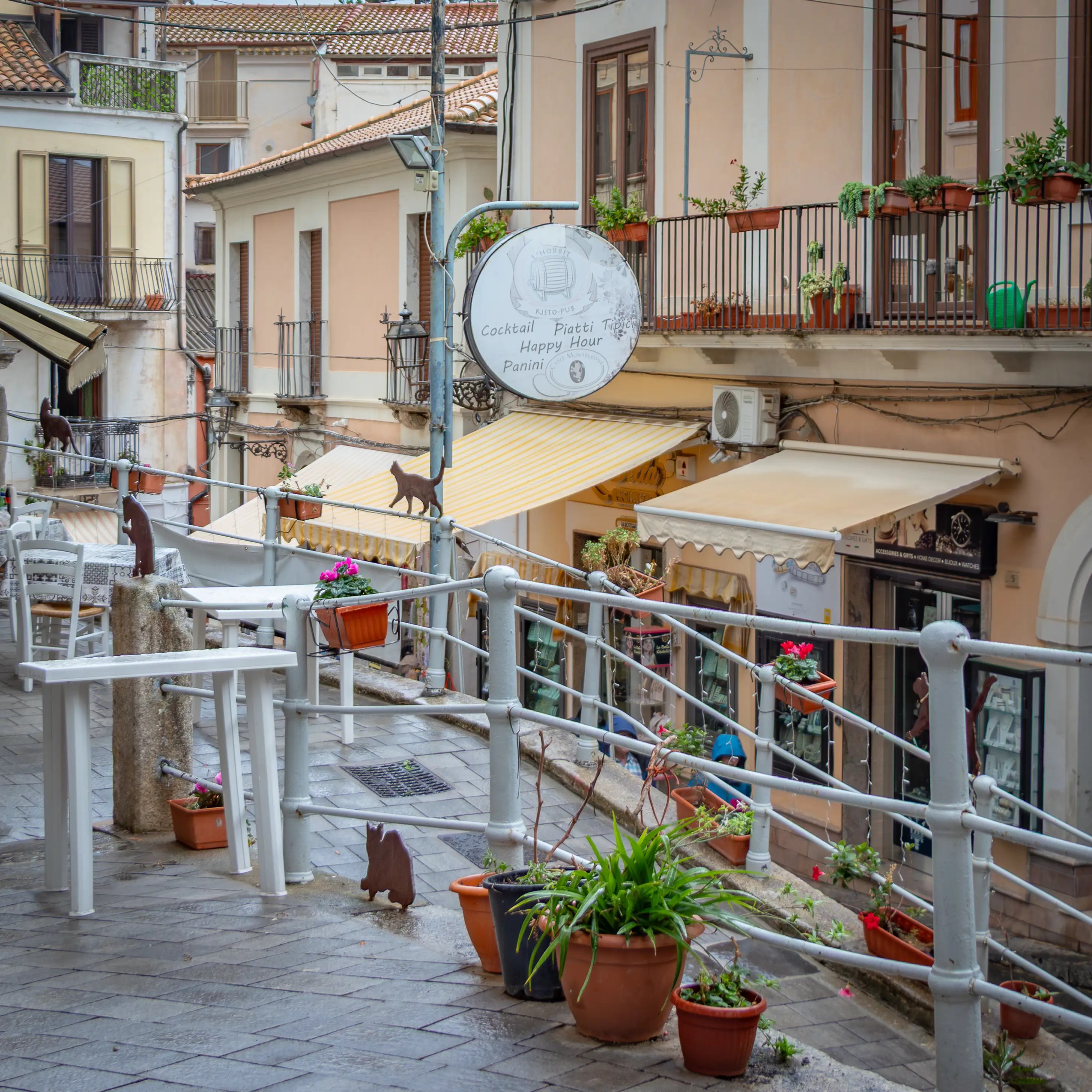 A raised walkway with metal railings, potted plants, and café signs overlooking a street lined with buildings and balconies.