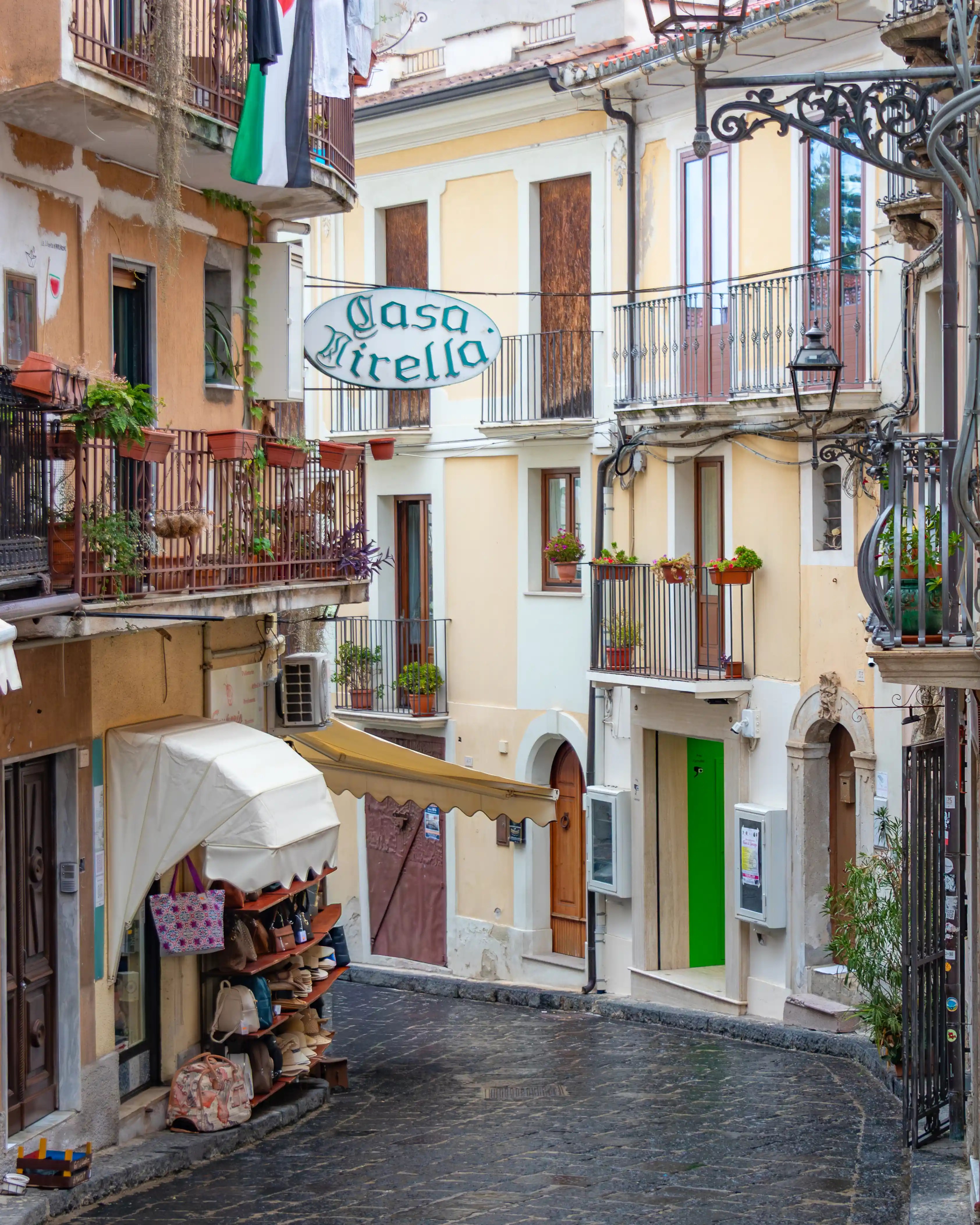 A narrow street with balconies, hanging plants, and a sign reading “Casa Mirella” above a small shop display.