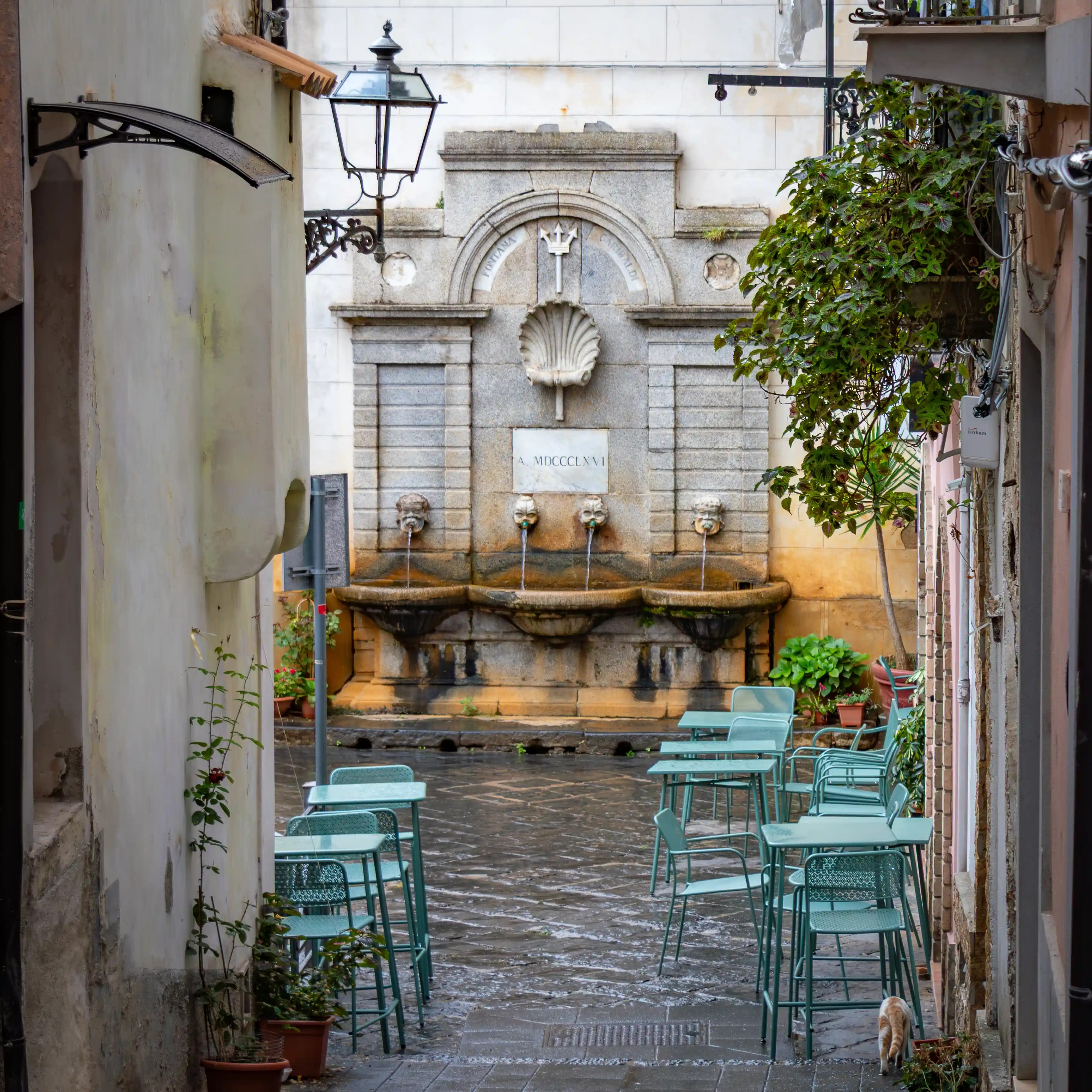An outdoor seating area with turquoise chairs and tables facing a stone fountain with multiple spouts in a small courtyard.