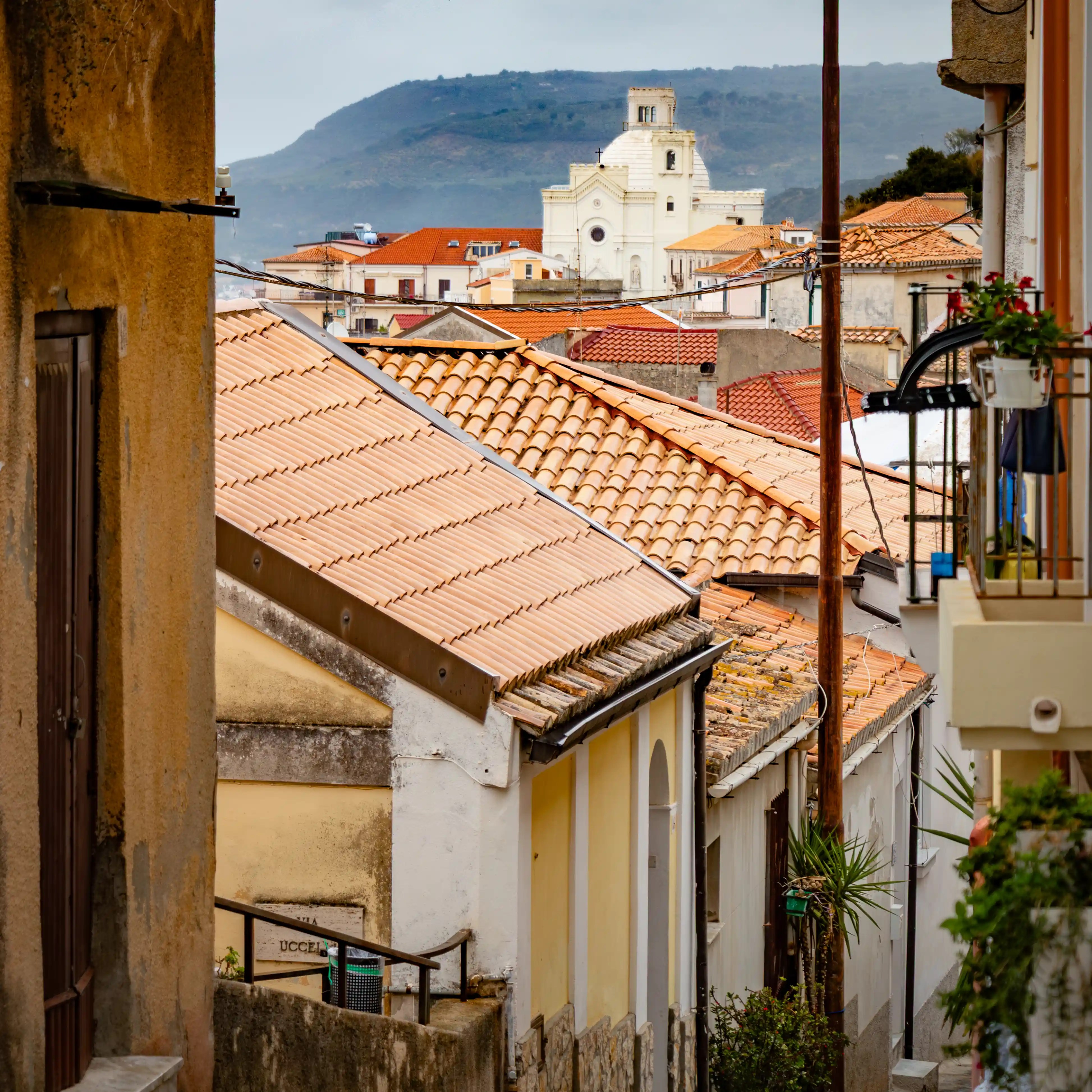 Terracotta rooftops and buildings in Pizzo with a white church visible in the distance against a hillside.