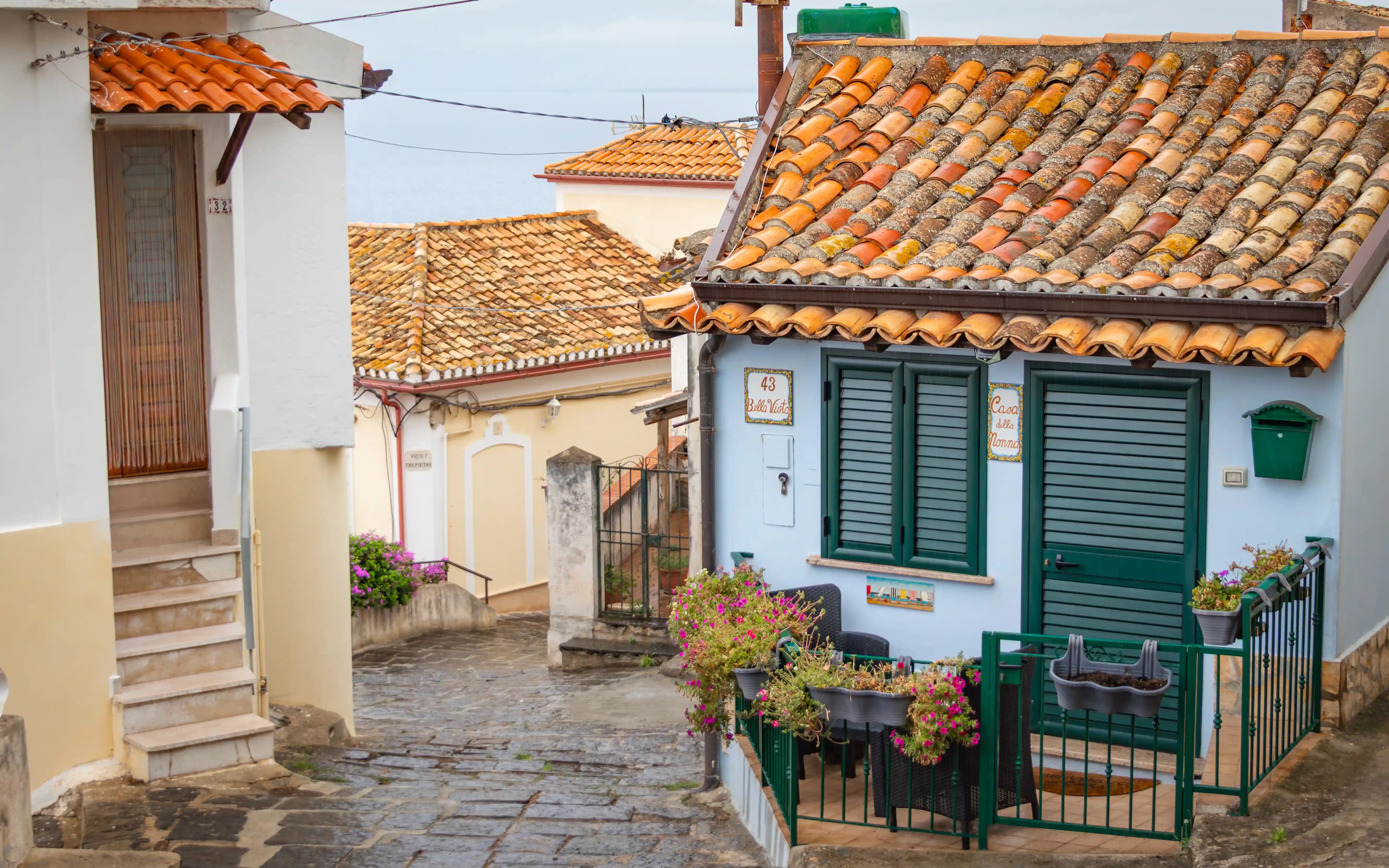 A small blue house with green shutters and a tiled roof surrounded by narrow streets and neighboring buildings.