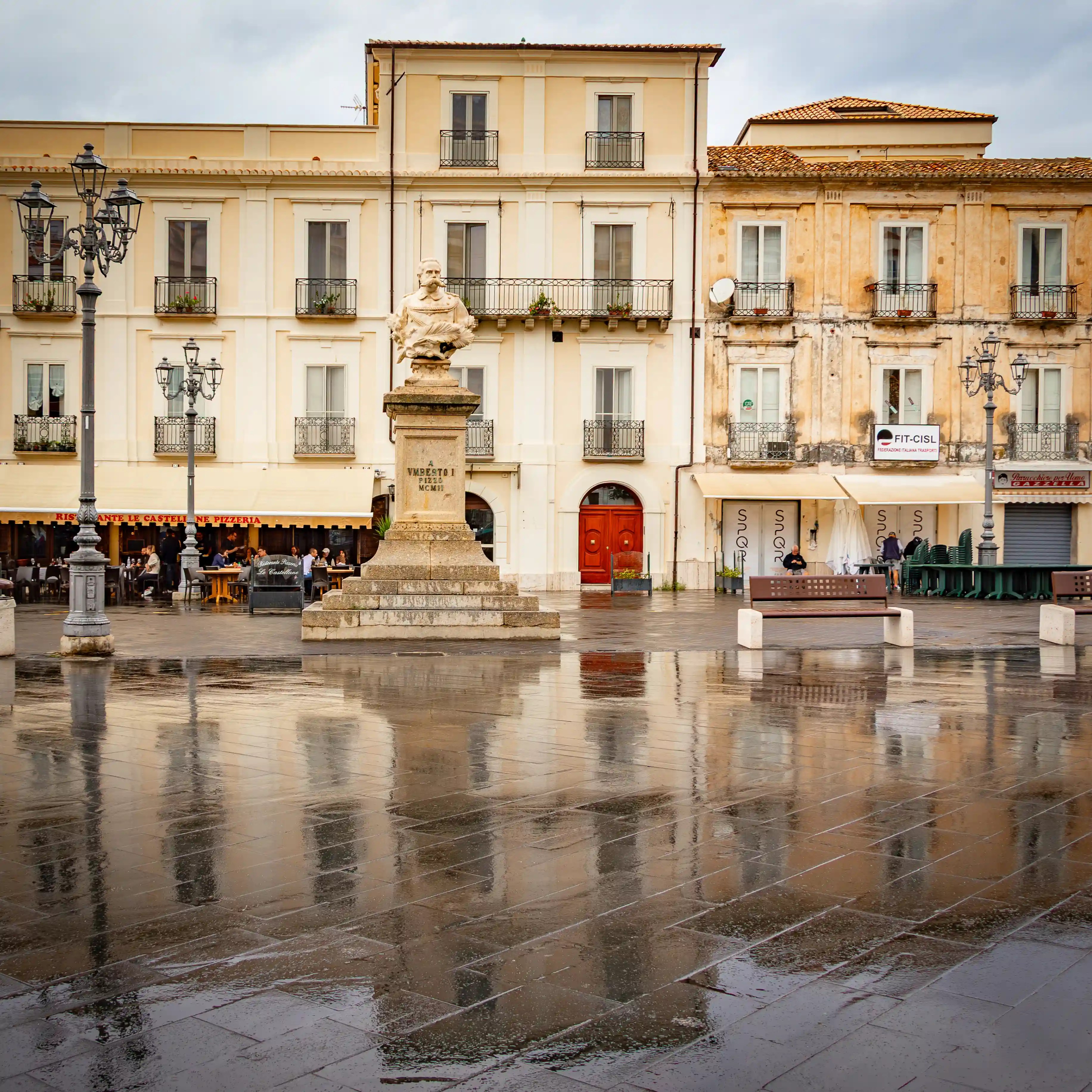 A statue on a pedestal stands in a large square with wet pavement reflecting surrounding buildings and street lamps.