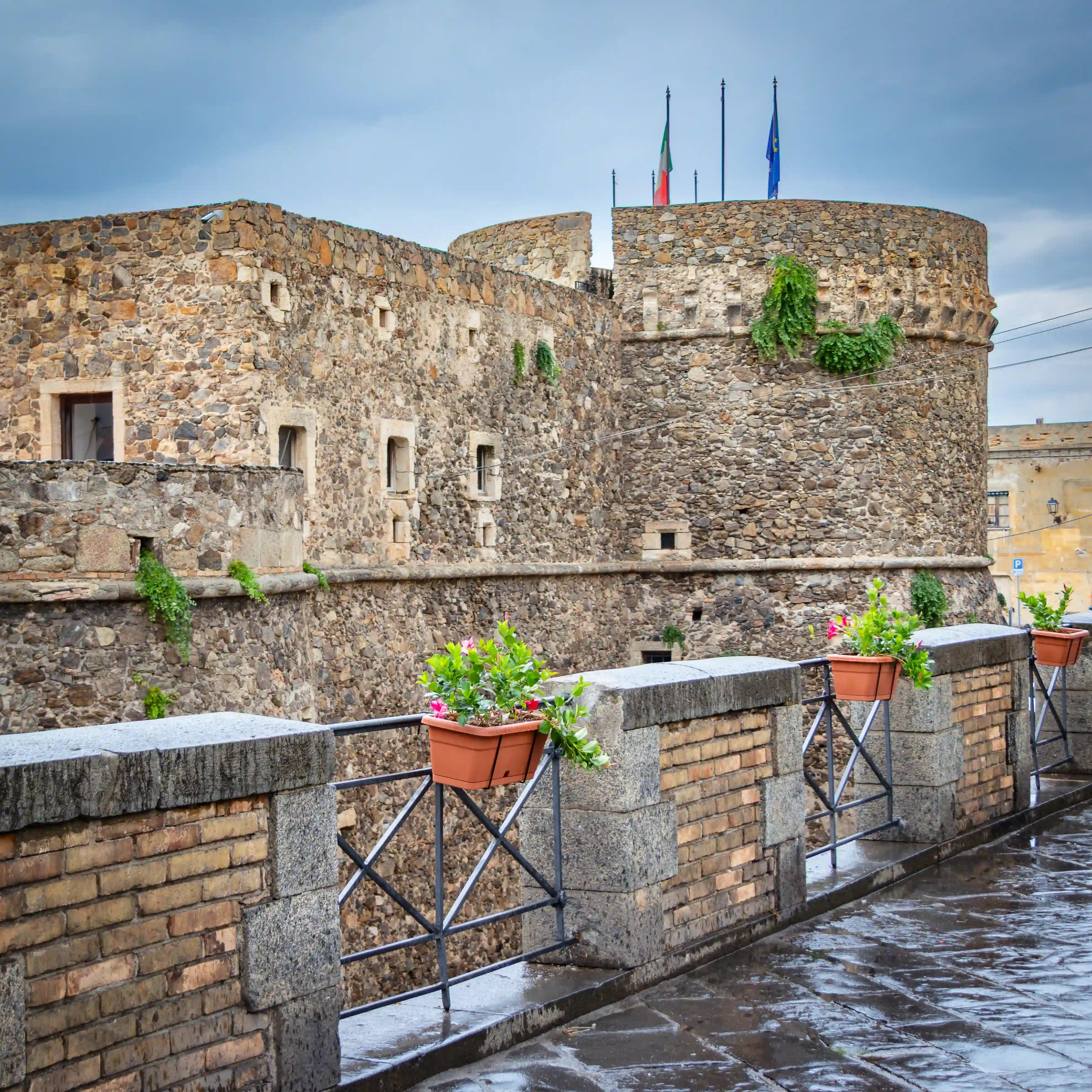 Stone fortress walls with small windows and a round tower topped with flags, viewed from a walkway with flower boxes.