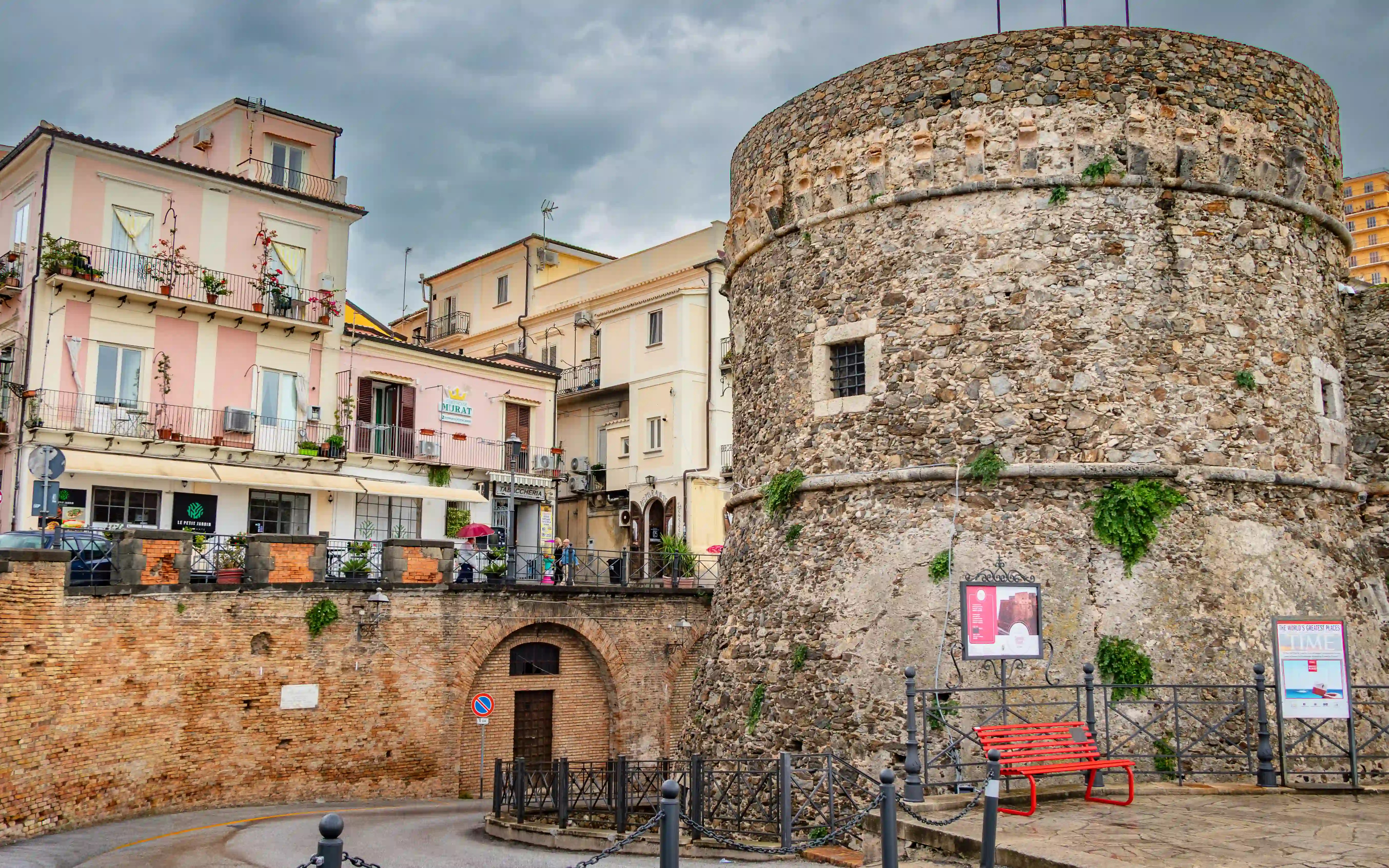 A round stone tower and adjacent wall of a fortress stand beside a street lined with buildings and balconies.