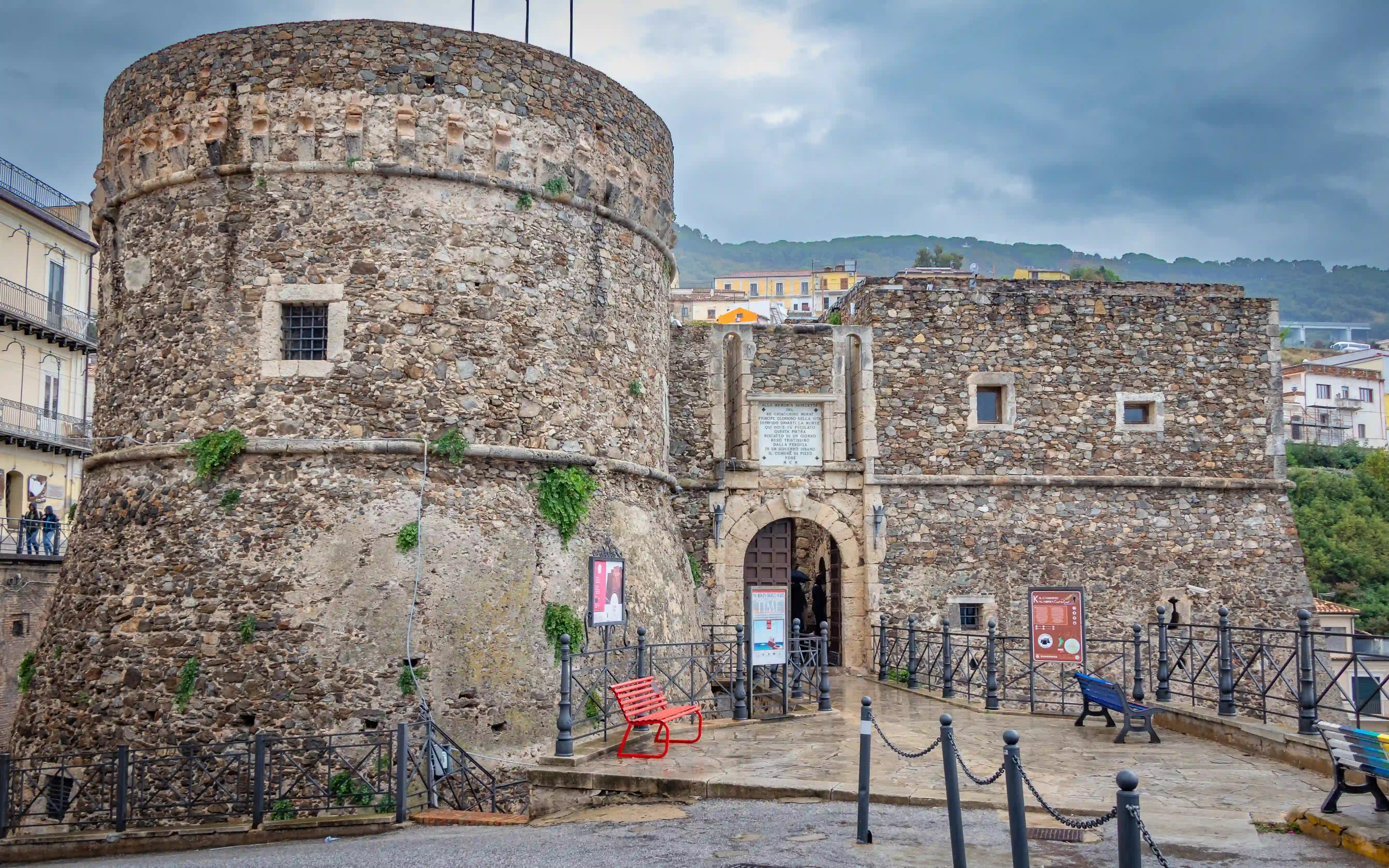 A stone fortress with a round tower and an arched entrance is accessed by a small paved area with railings and benches.
