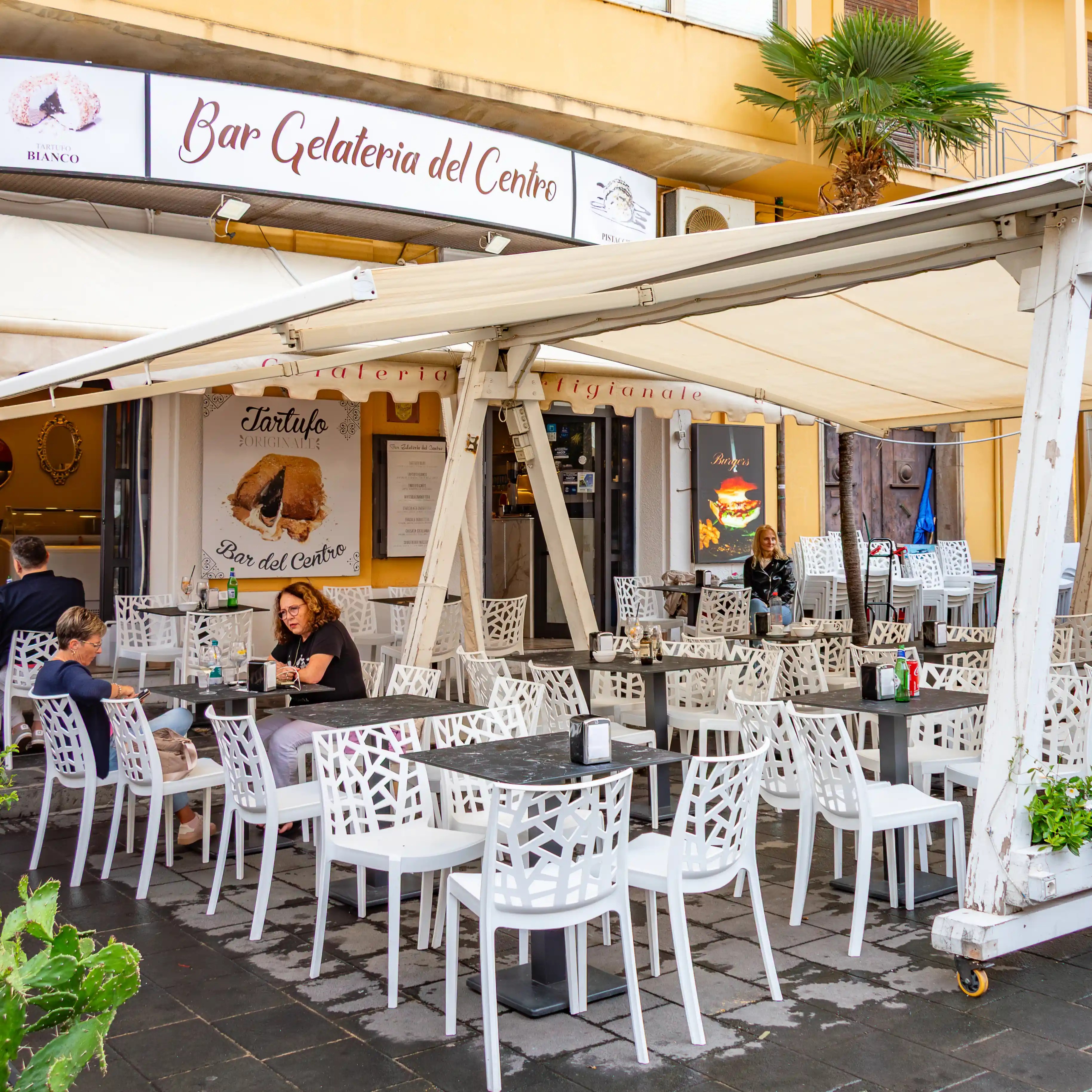 Outdoor café seating with white chairs and tables sits beneath awnings in front of Bar Gelateria del Centro.
