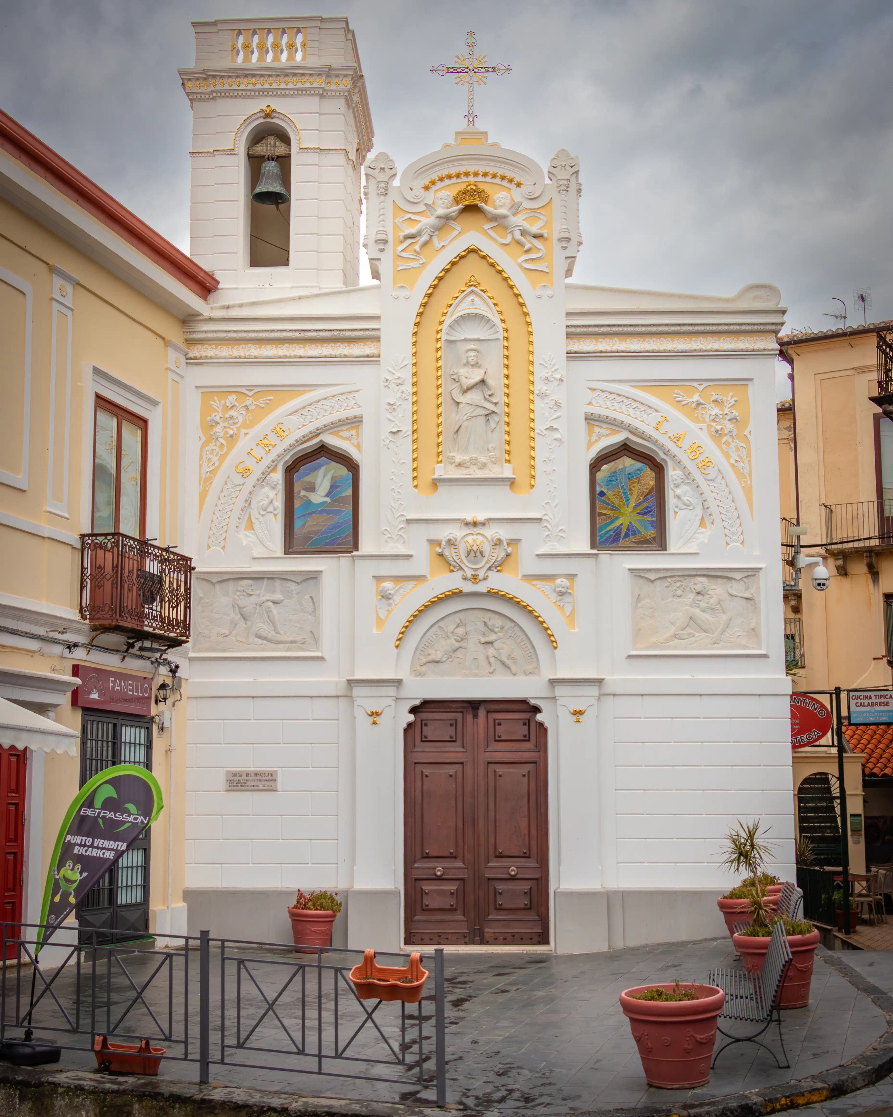A white church façade with gold detailing, statues, and a bell tower stands along a narrow street.
