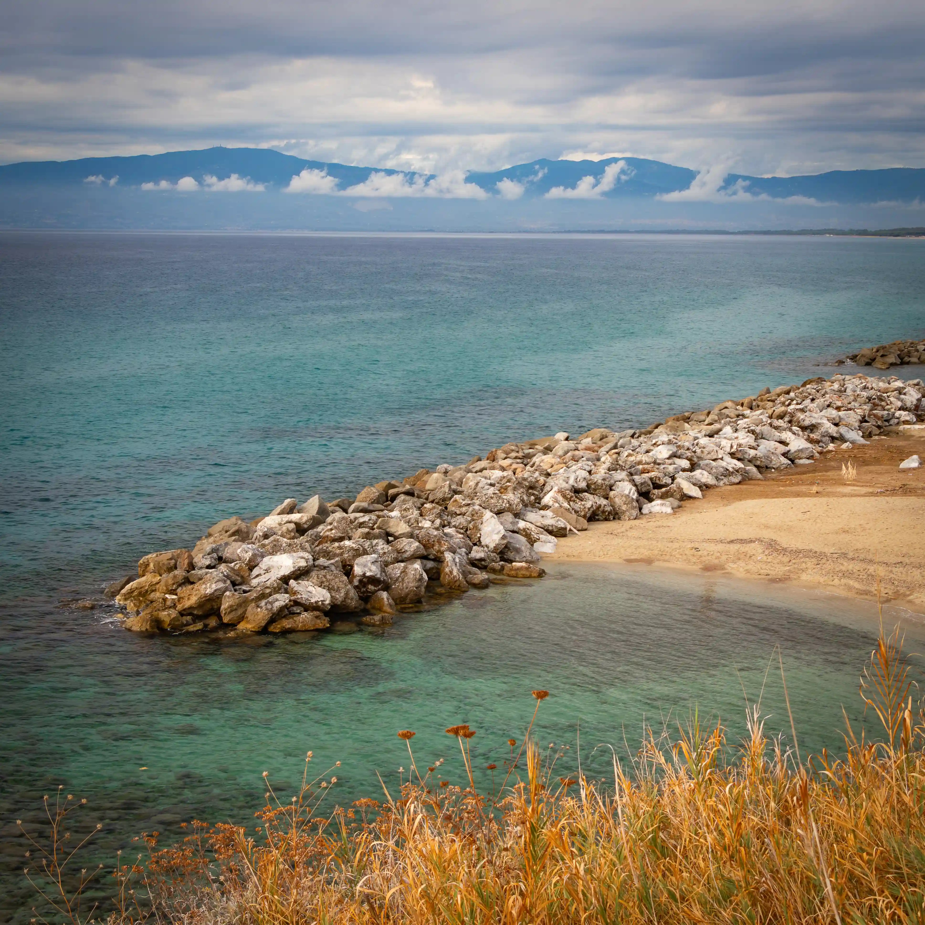 Rock breakwater curving into clear turquoise water with distant mountains under low clouds.