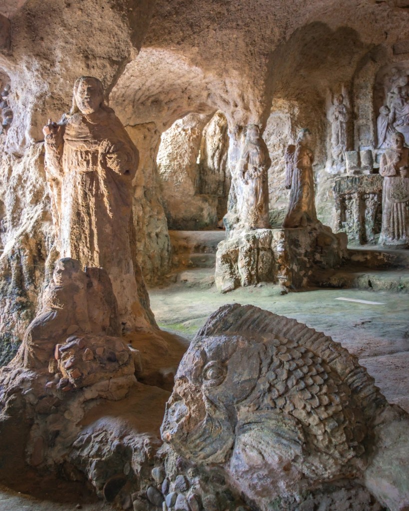 Carved stone figures and religious scenes inside a cave with rough rock walls and arched openings.