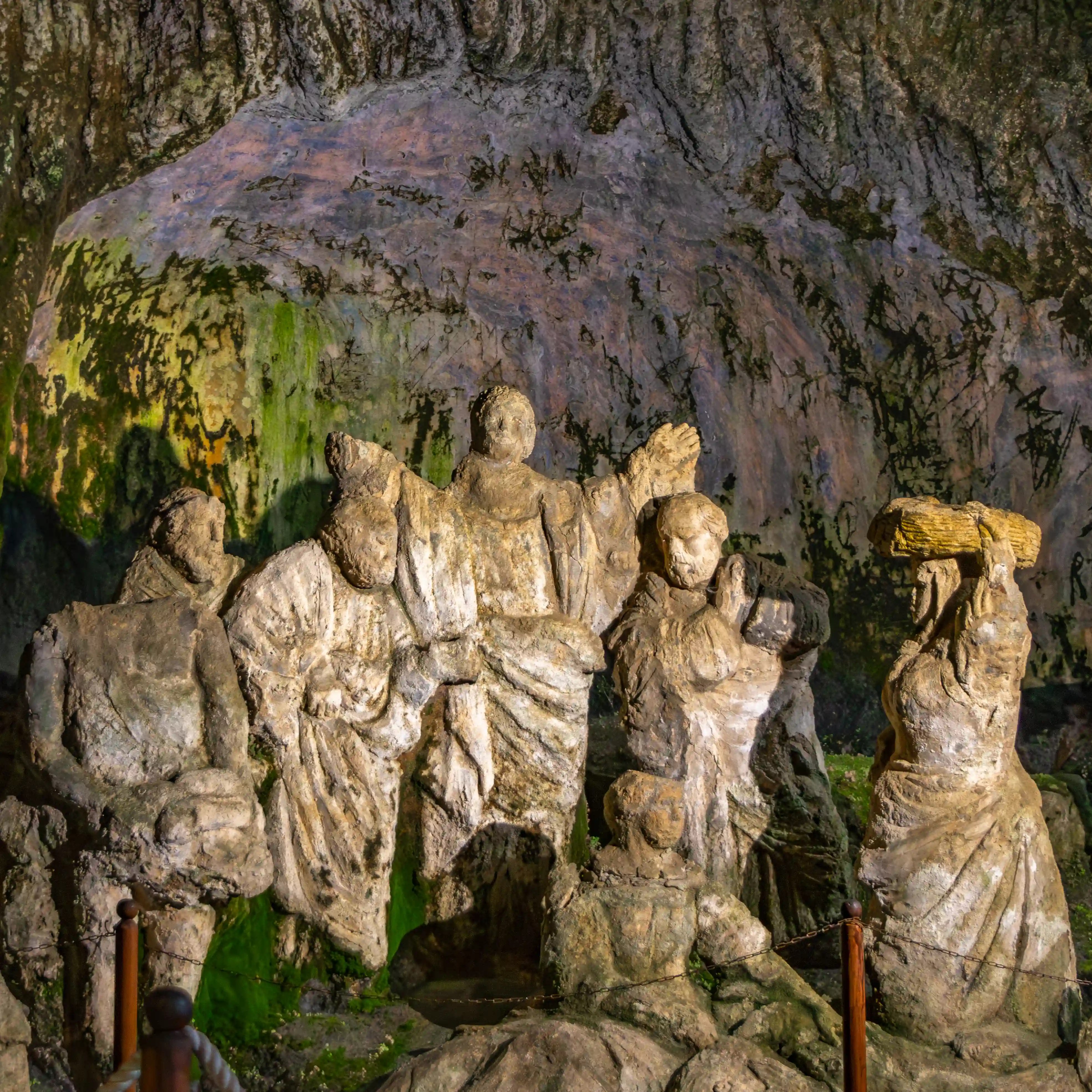 Stone statues of multiple figures arranged in a cave setting with green mineral staining on the rock.