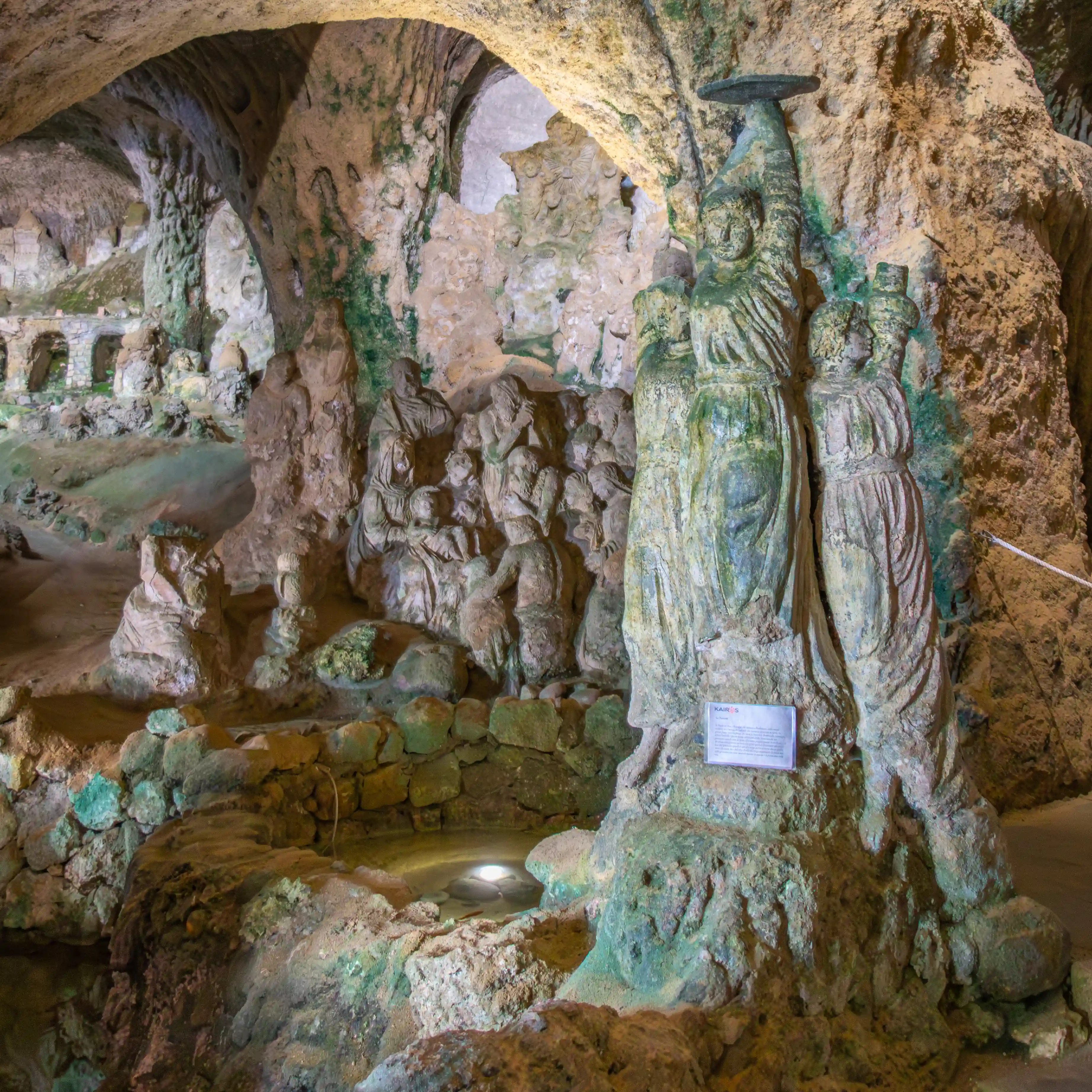 Carved statues beneath a rock arch inside a cave with a small pool of water in the foreground.