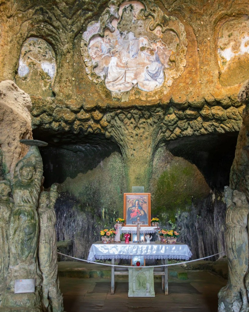 Small altar with religious icon and flowers set within a cave framed by carved stone figures.