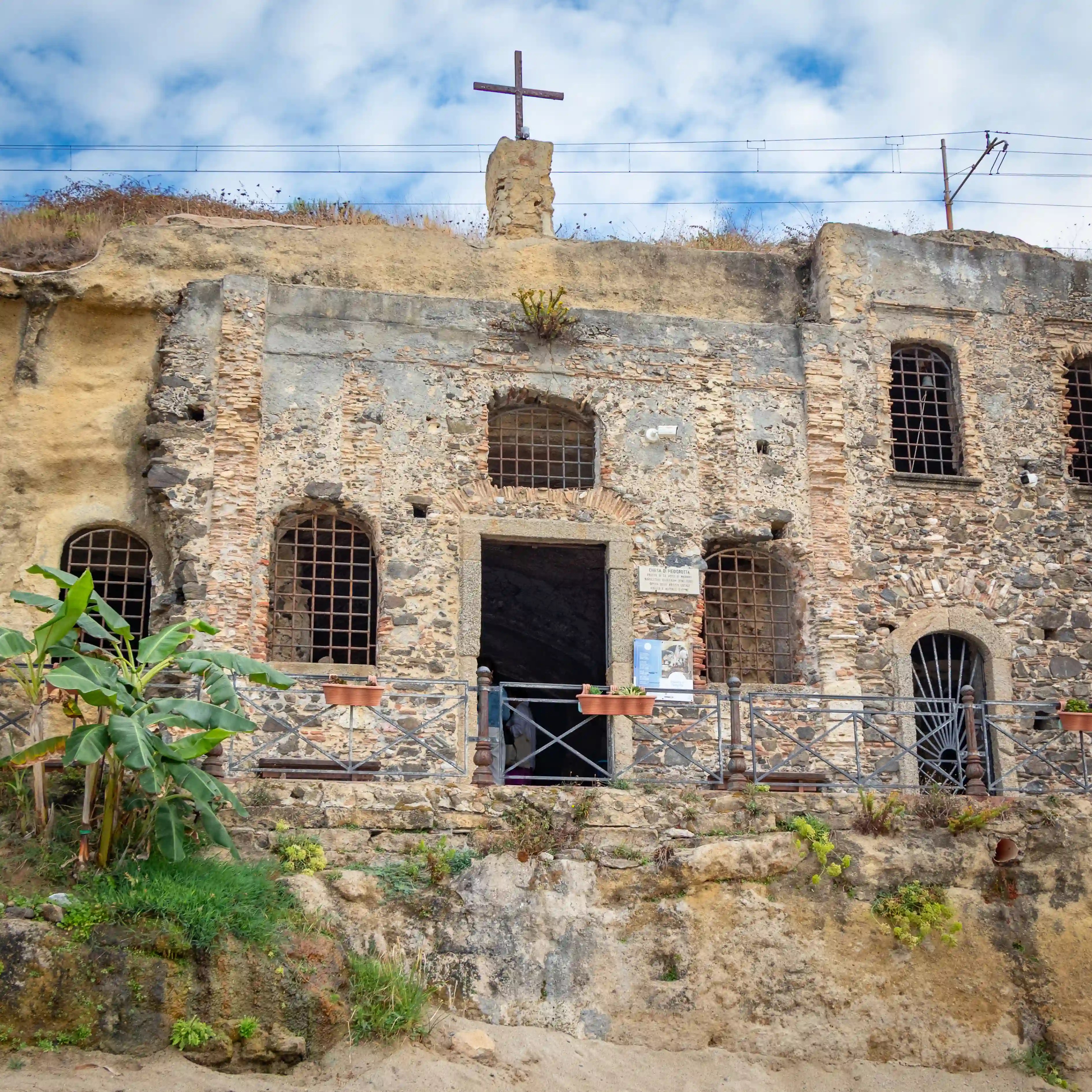 Stone building built into a hillside with arched windows, a central doorway, and a cross on top.