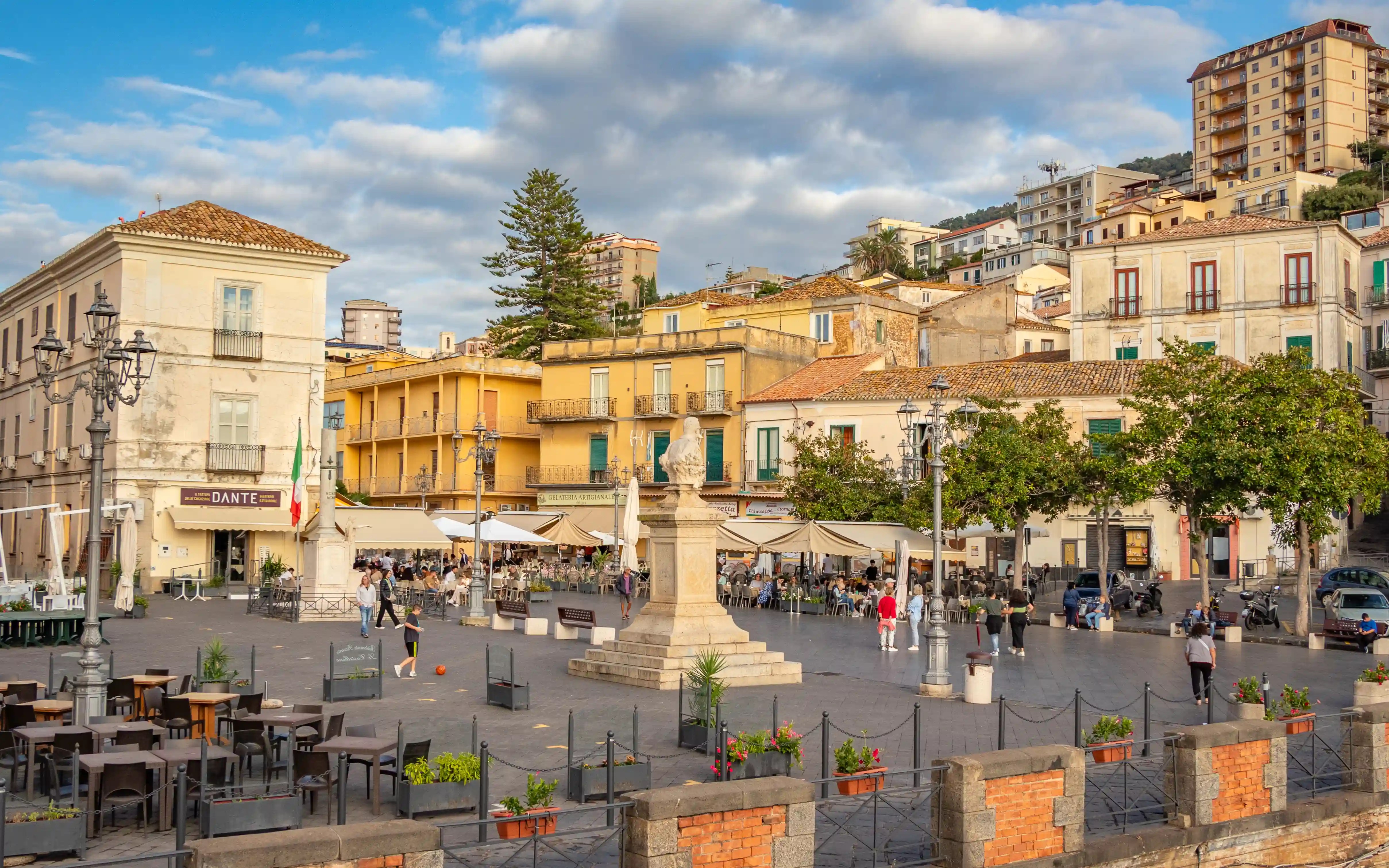 A large square with people walking, outdoor seating, and a central statue is surrounded by colorful buildings.