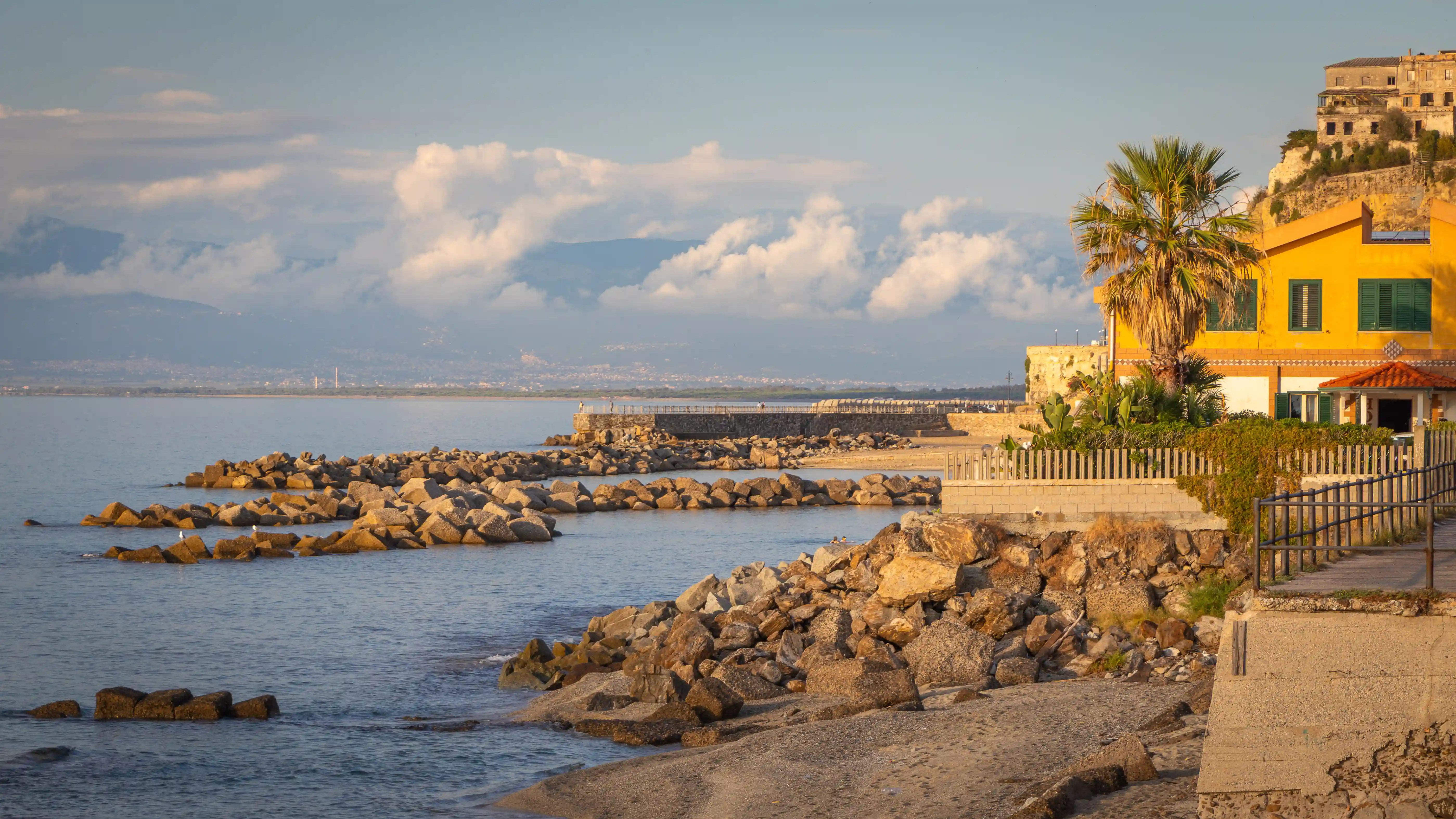 A coastal scene with rocky breakwaters, calm water, and a yellow building with a palm tree along the shoreline.