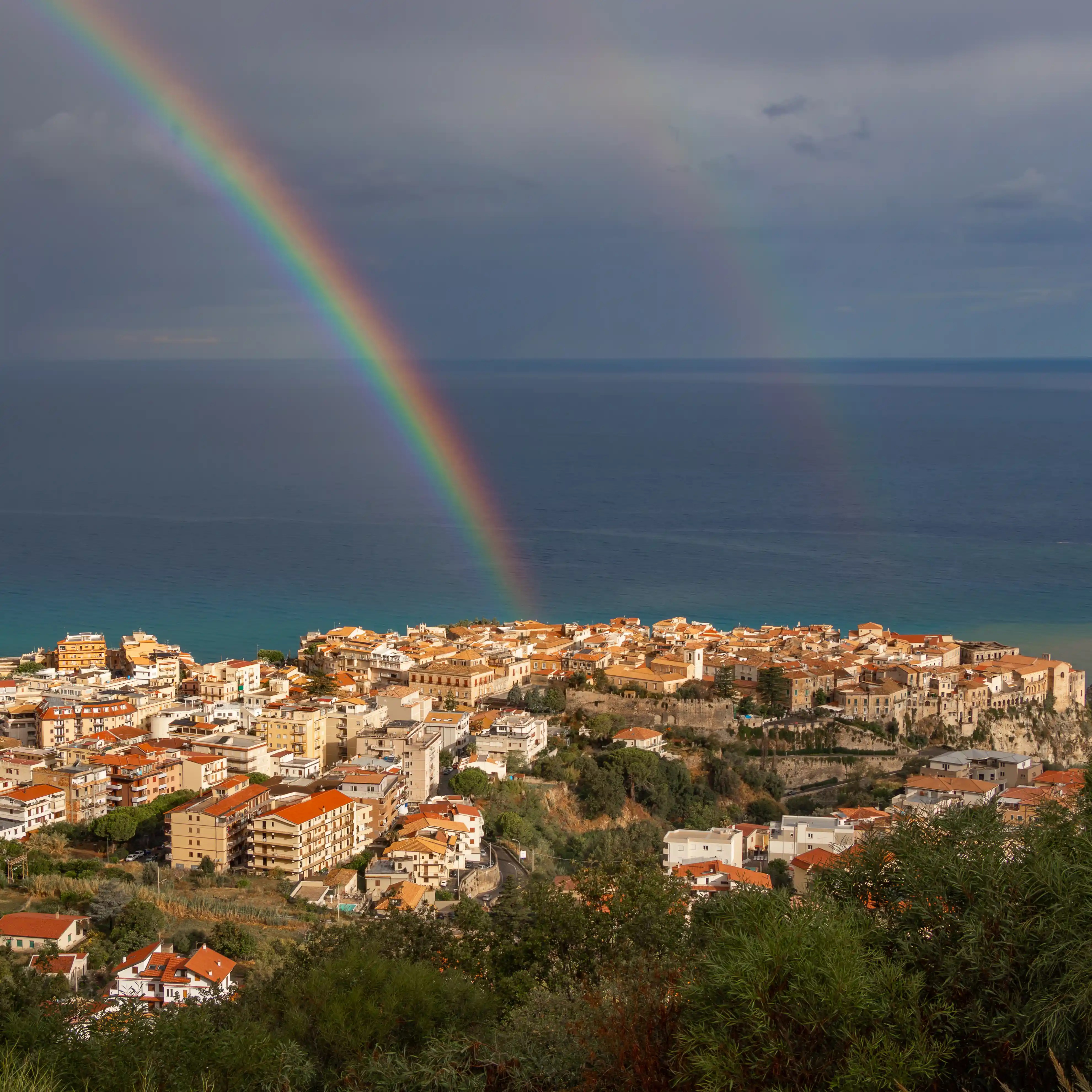 View of Tropea’s old town on a cliff with a bright rainbow over the sea in the background.