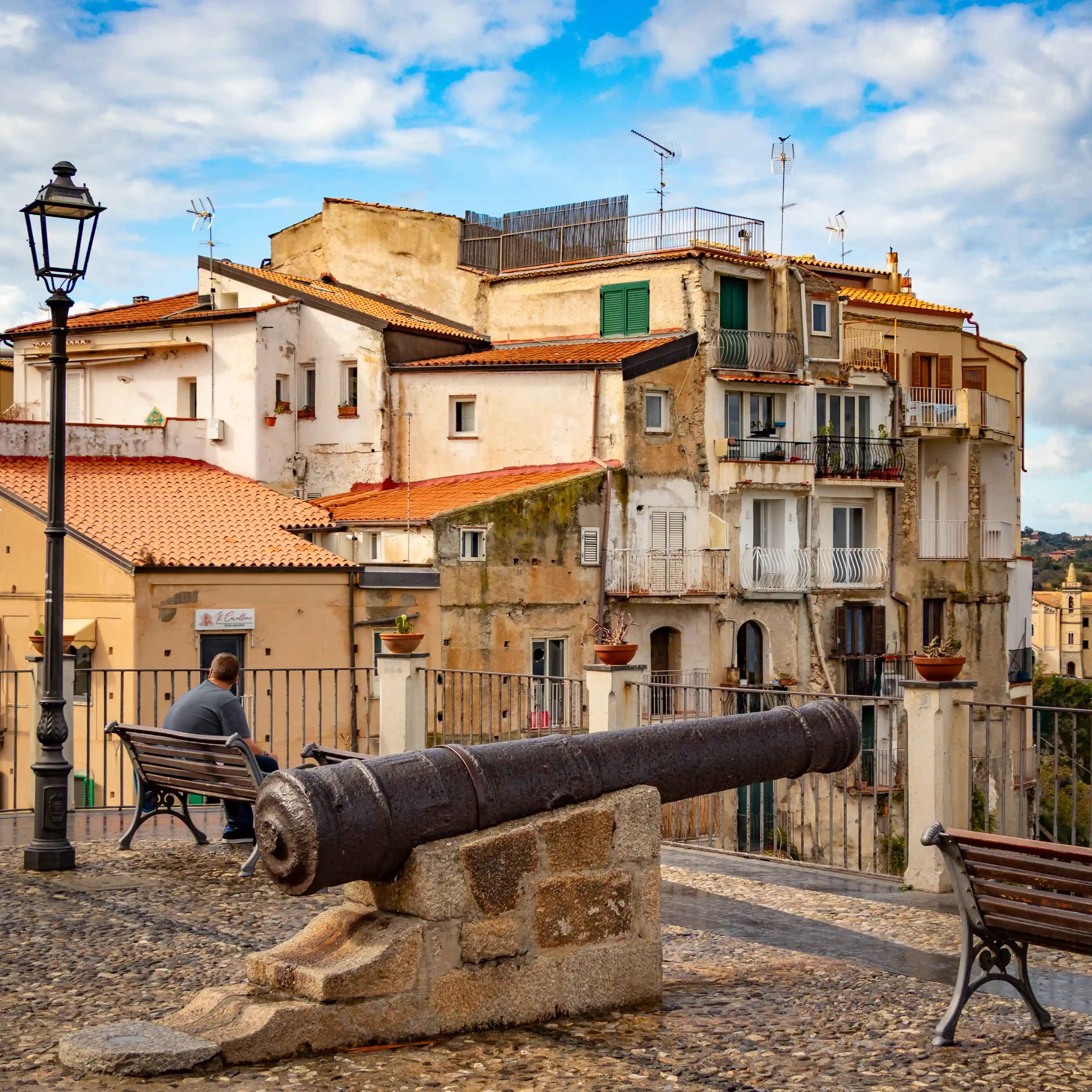 Old cannon mounted on a stone base near benches overlooking buildings in Tropea.