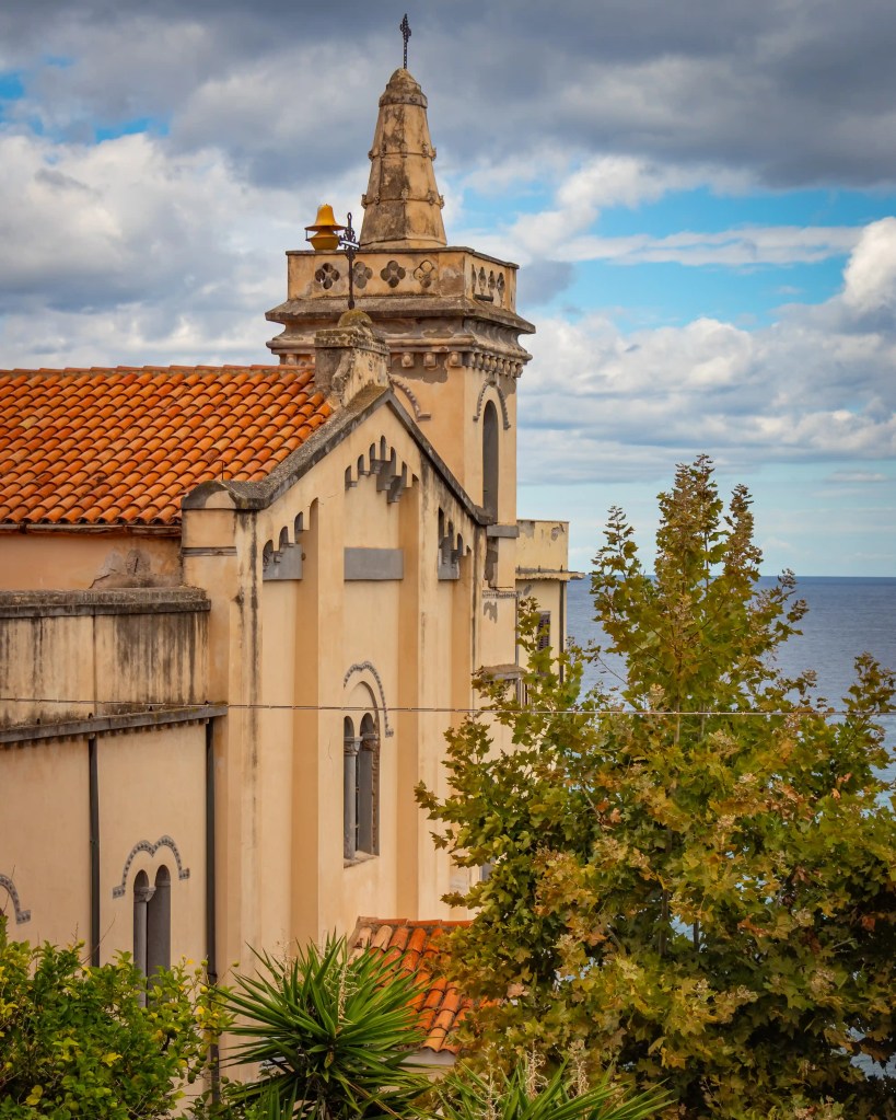 Church building with a bell tower and tiled roof partially framed by trees with the sea behind it.