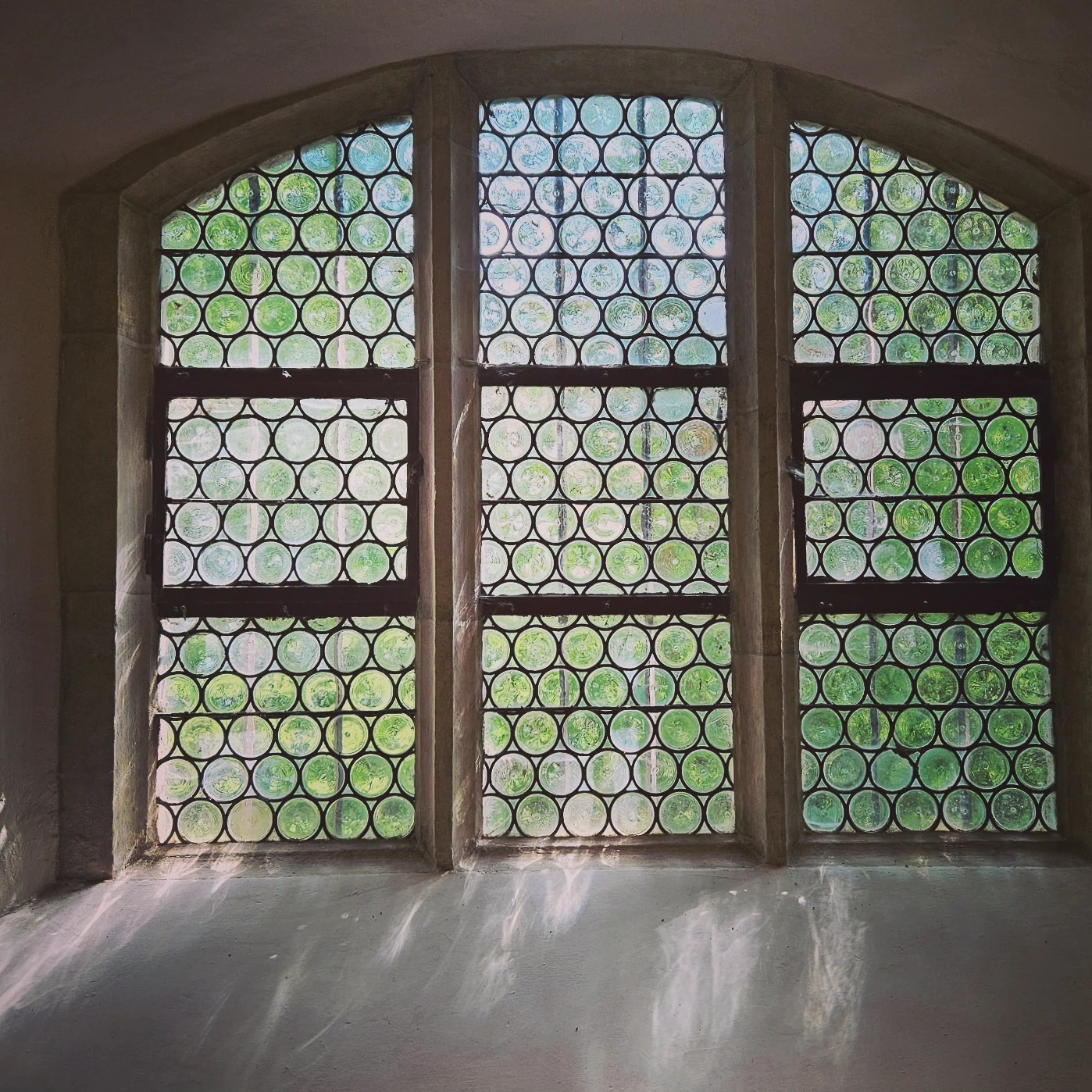 A three-panel window filled with circular glass panes casts soft light onto a white interior wall.