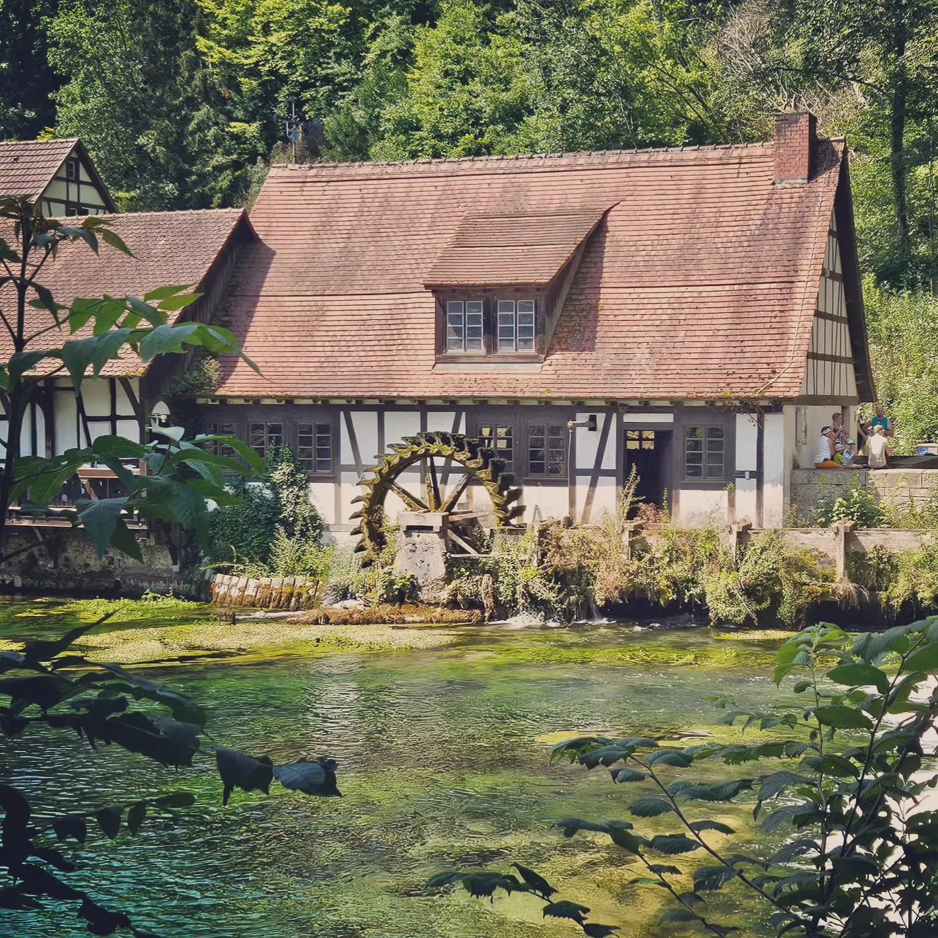 The half-timbered Blautopf mill and water wheel sit along the clear stream, framed by leafy branches in the foreground.