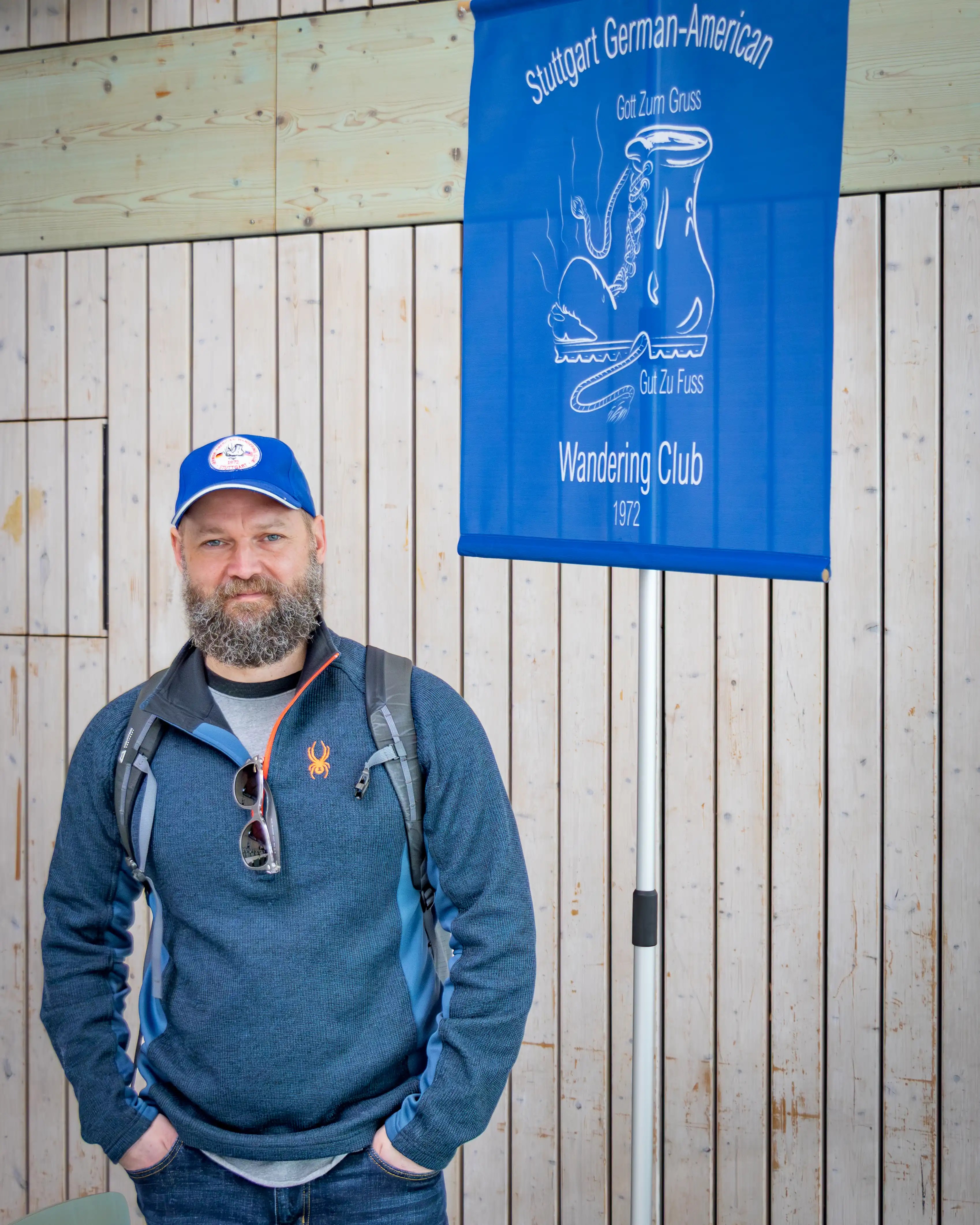 A man wearing a blue cap and backpack stands beside a blue banner reading “Stuttgart German-American Wandering Club.”
