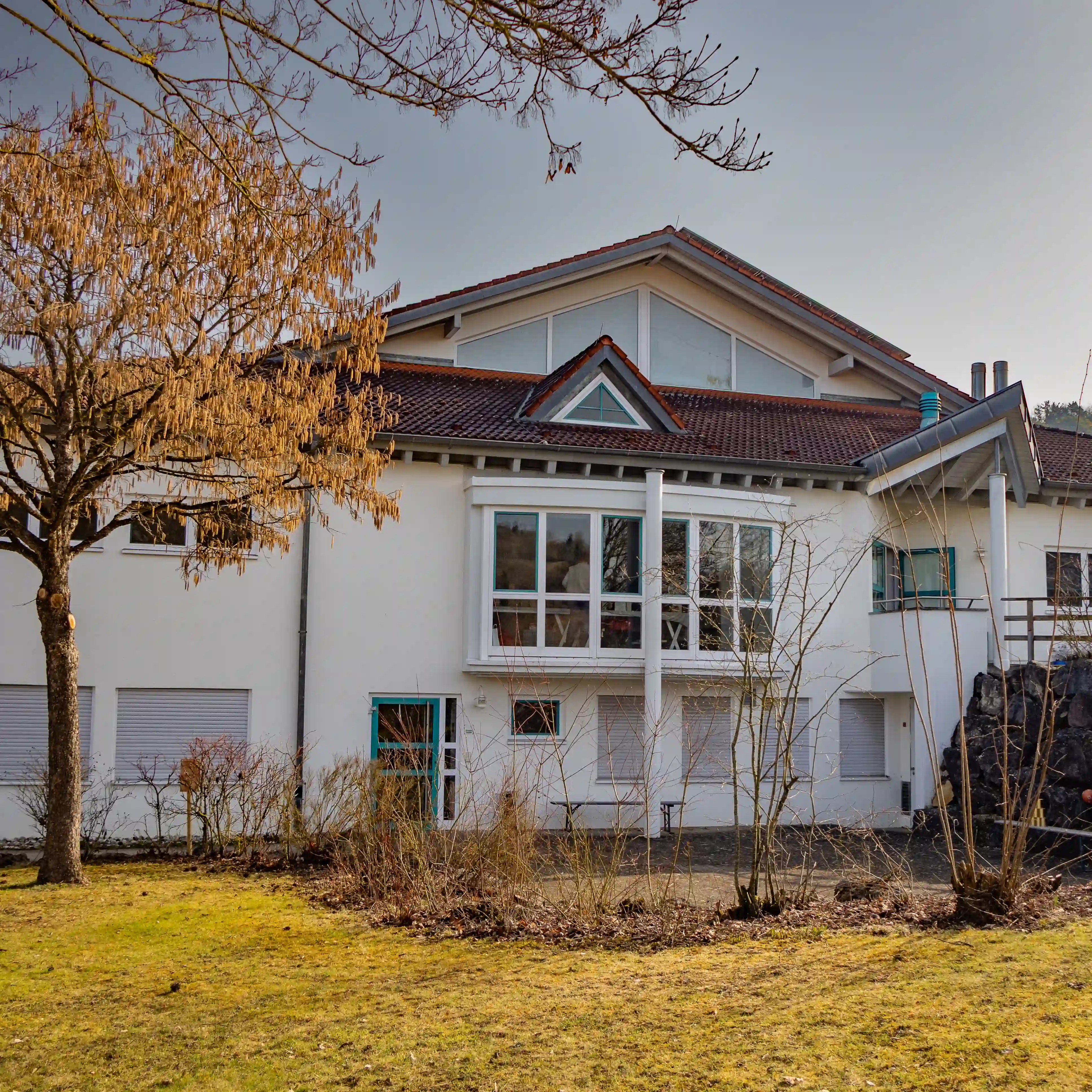Light-colored building with a red tiled roof and large windows surrounded by trees and grass in Lauterach, Germany.
