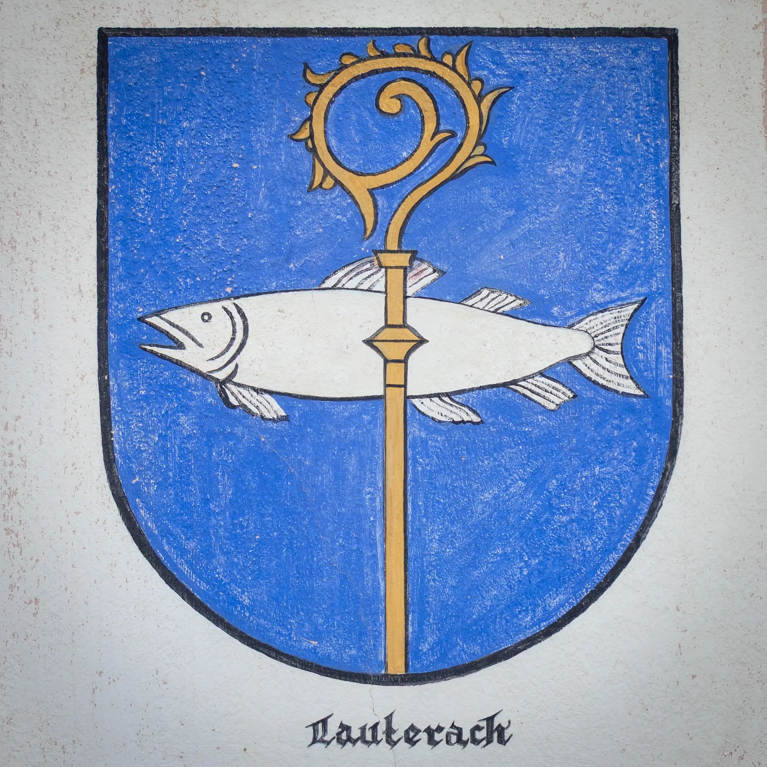 A blue shield displaying a white fish crossed by a vertical gold staff with the word “Lauterach” written below.