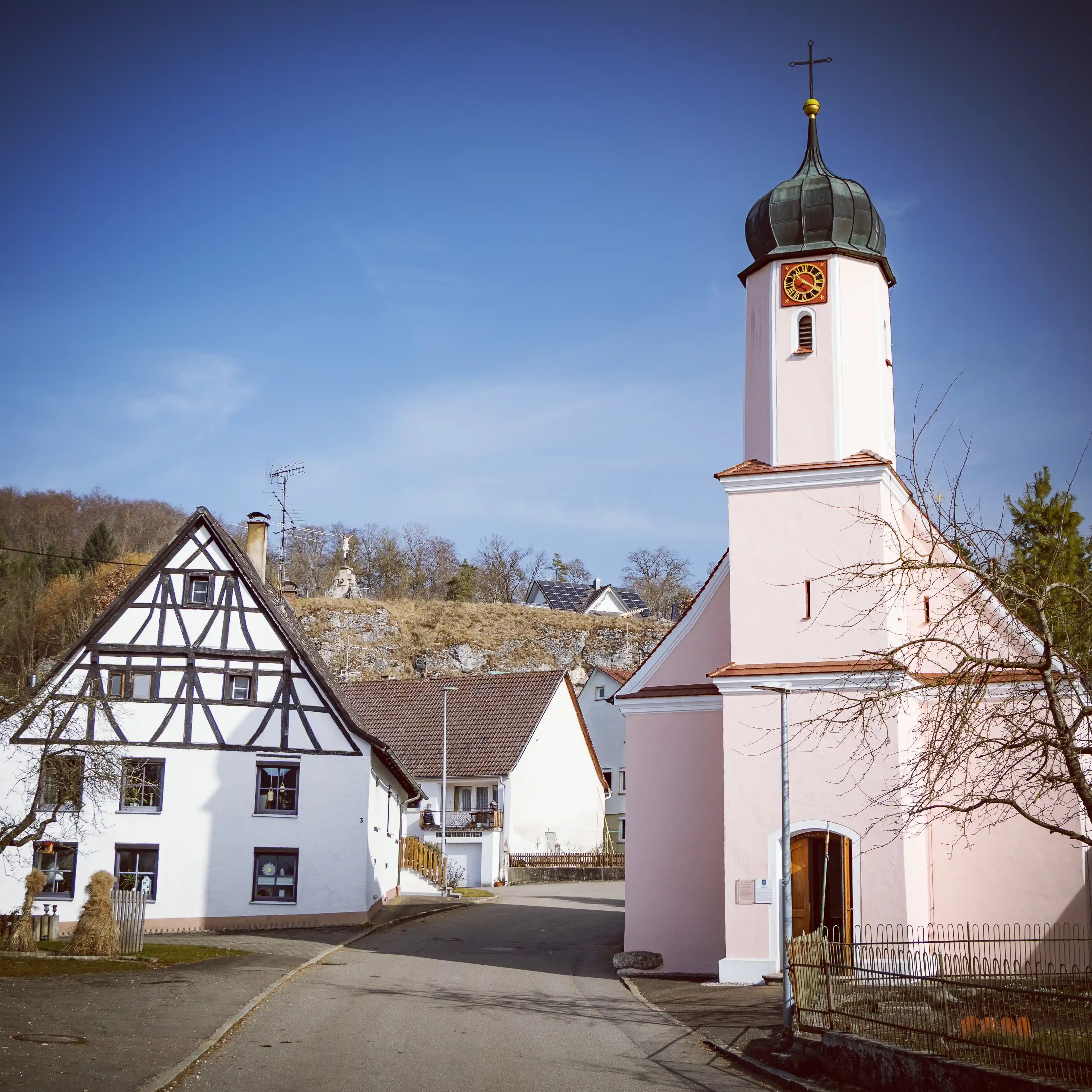 A small church with a pale pink exterior and onion-domed tower stands beside a half-timbered house along a quiet village street.