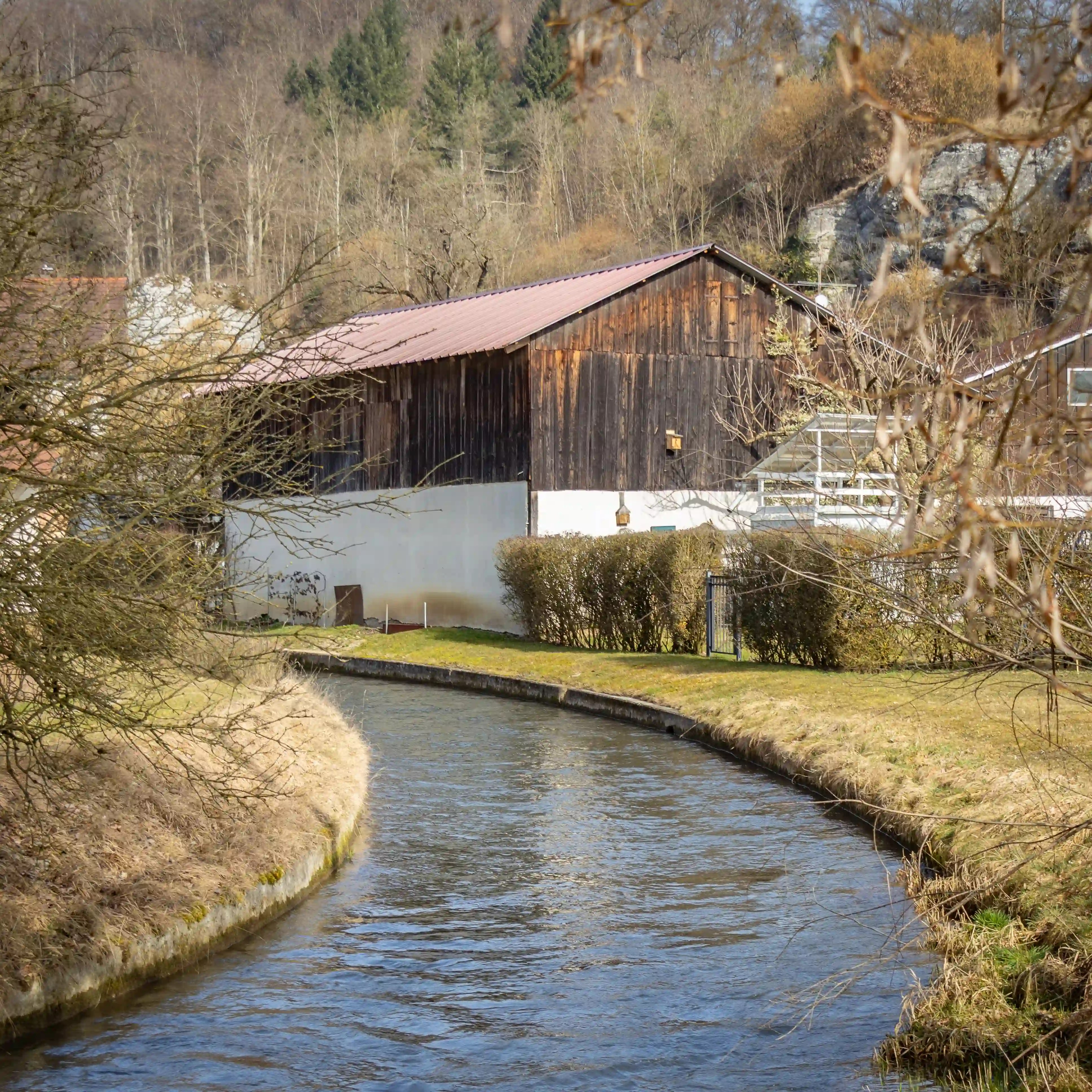 A narrow stream flowing past a wooden building and shrubs with trees and limestone cliffs in the background.