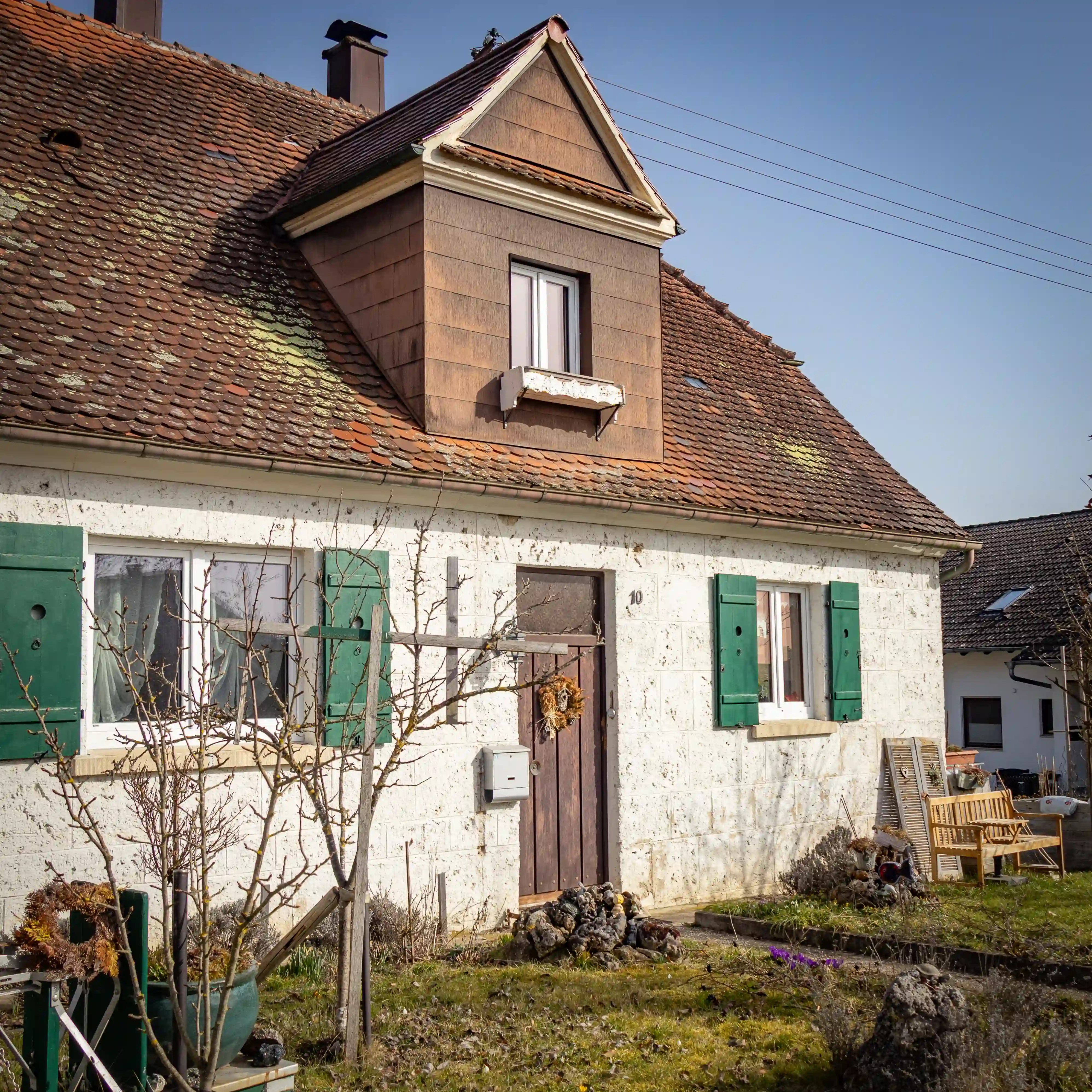A small house with green shutters and a steep tiled roof surrounded by a garden and yard.