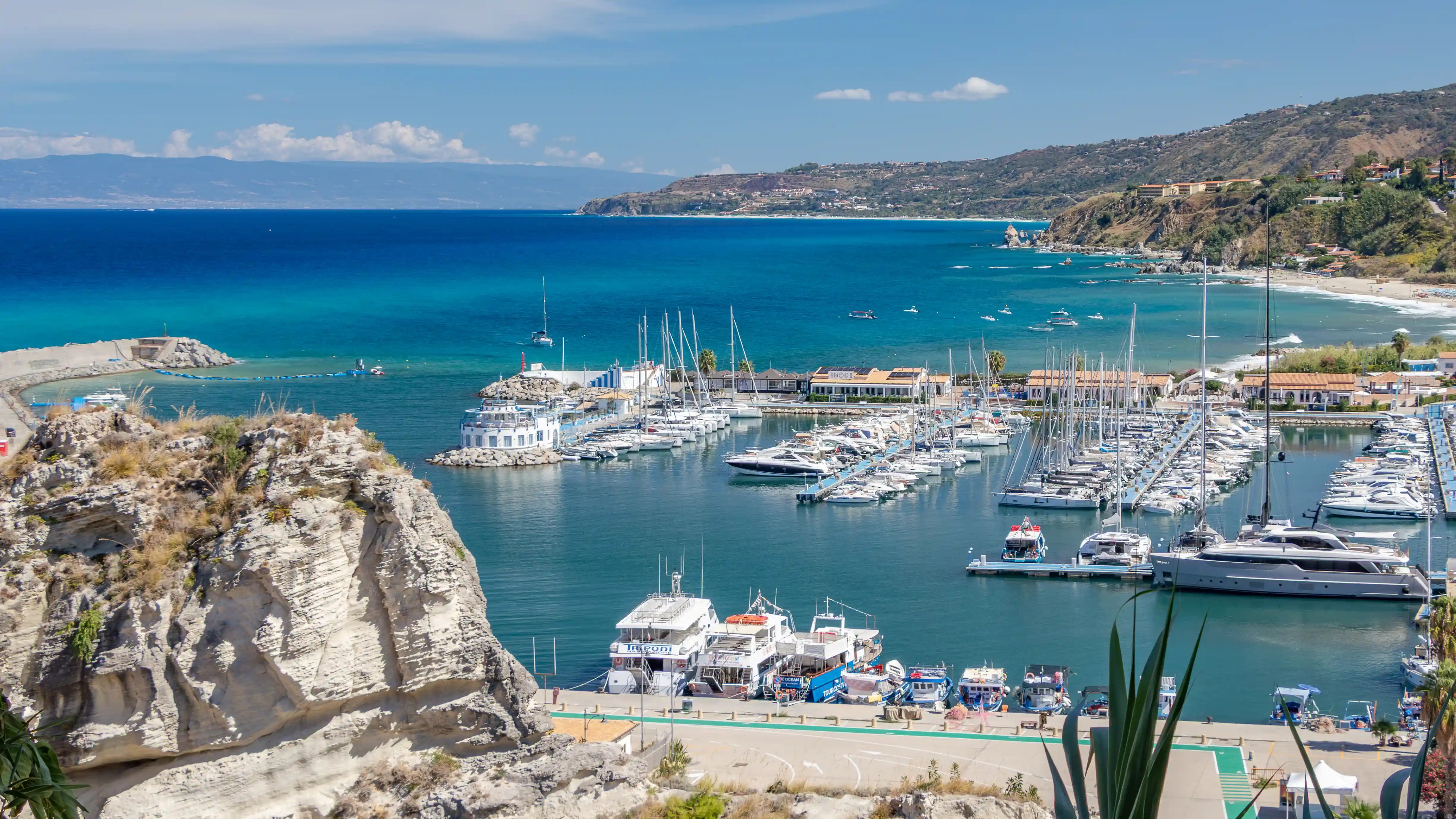 A marina filled with sailboats and yachts beside a rocky cliff and turquoise sea along a coastal town.