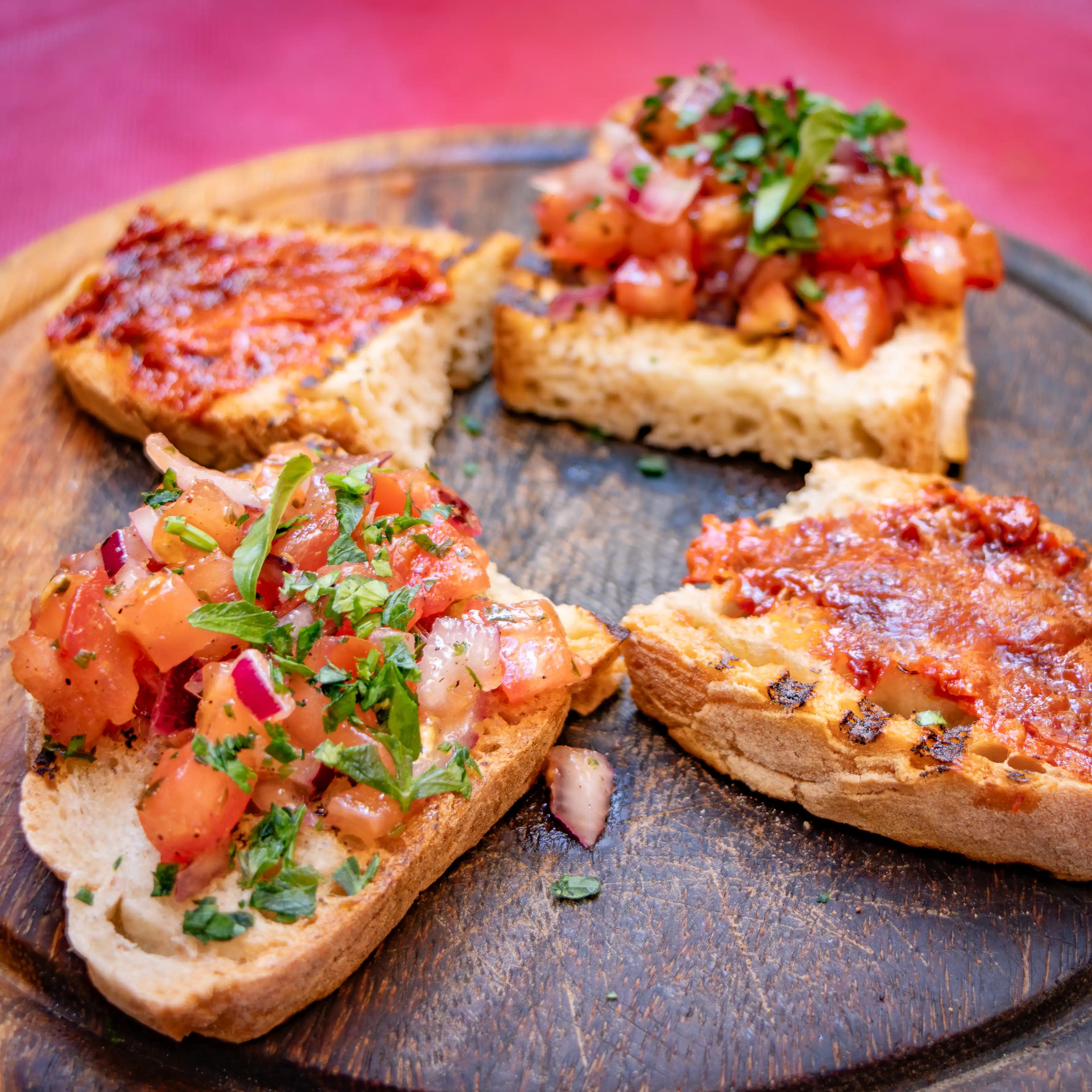 Four pieces of toasted bread topped with chopped tomatoes, herbs, and a red spread on a wooden plate.