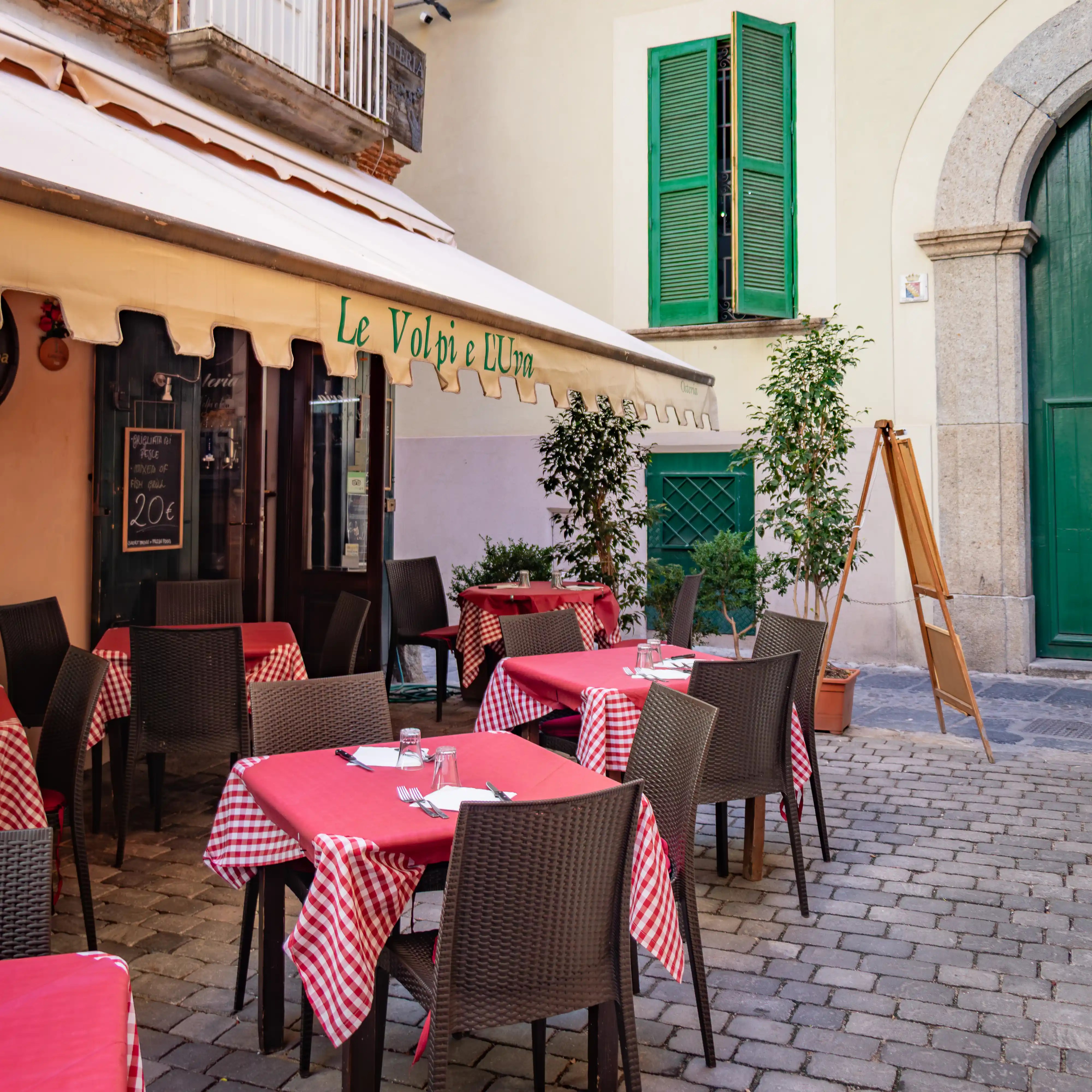 Outdoor seating area with red checkered tablecloths in front of a small restaurant with a striped awning and green shutters.