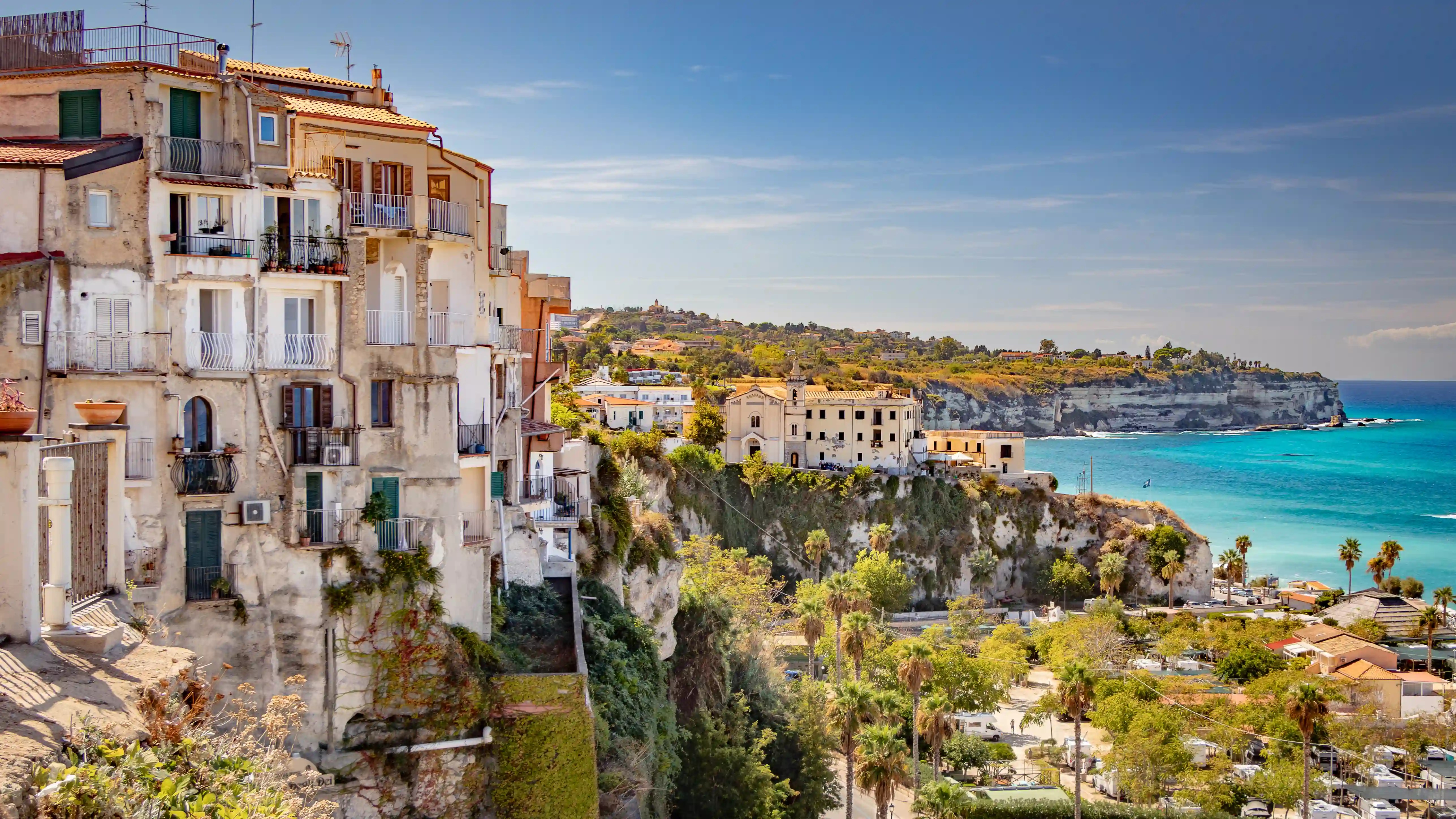 View of Tropea’s coastline with buildings on cliffs above turquoise water and a sandy beach below.