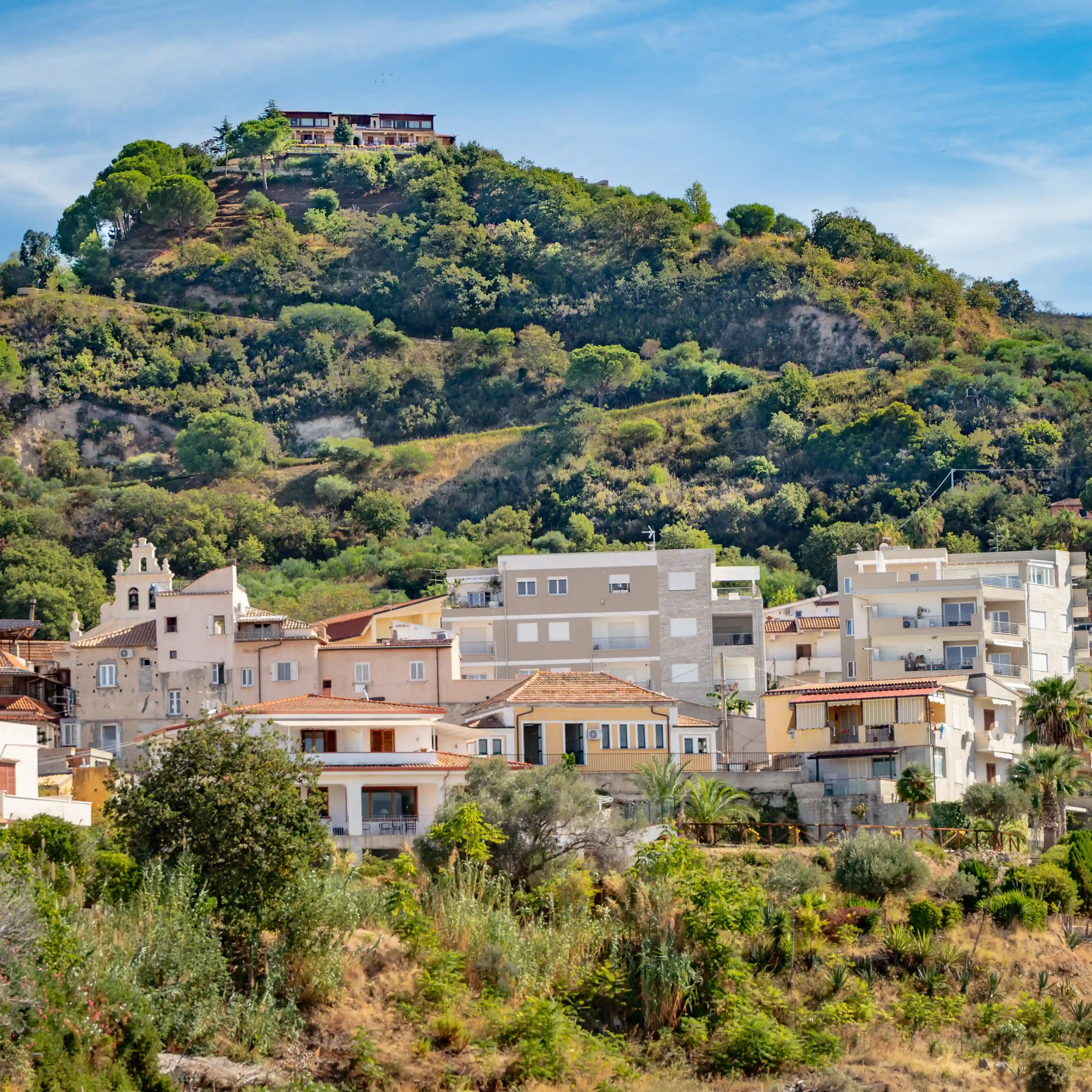 Cluster of houses at the base of a green hillside with a larger building at the top.