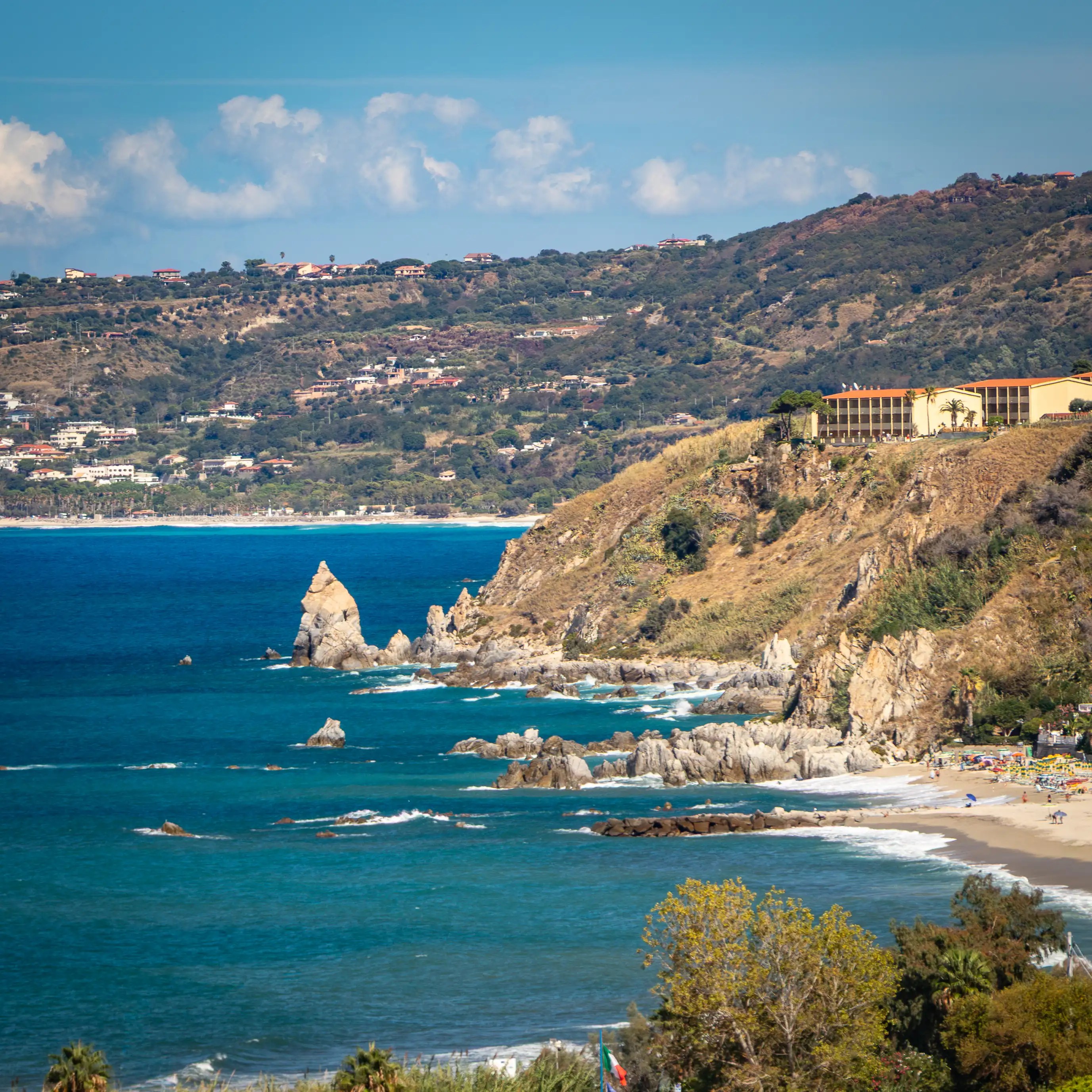 Rocky coastline with cliffs, blue water, and a sandy beach with umbrellas.