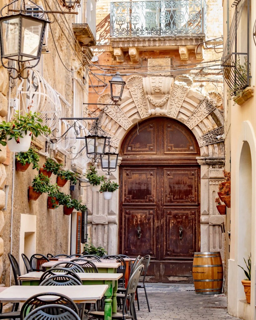 Outdoor café tables and chairs in front of a large arched wooden doorway with carved stone details.