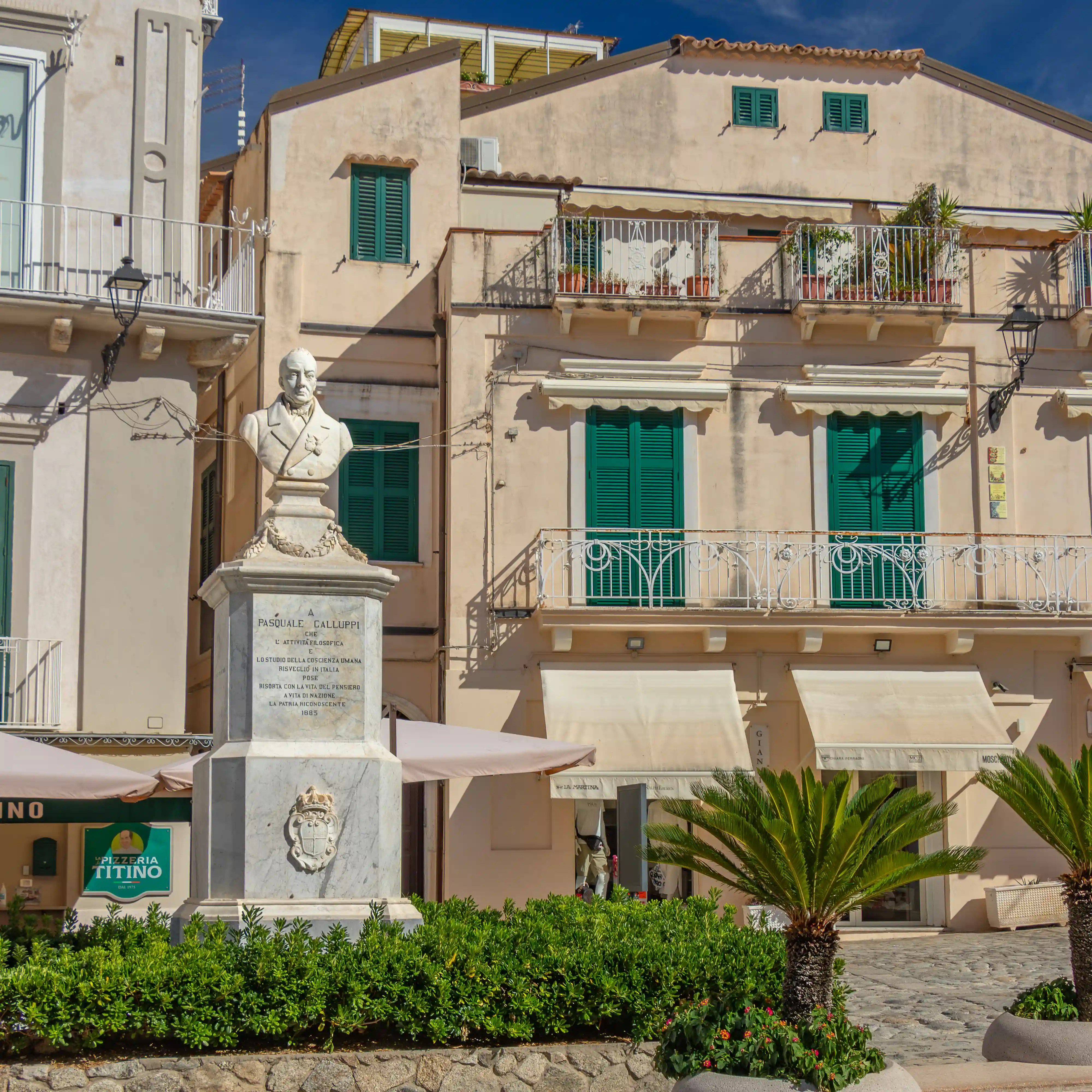 Bust statue on a pedestal in front of buildings with green shutters and balconies.
