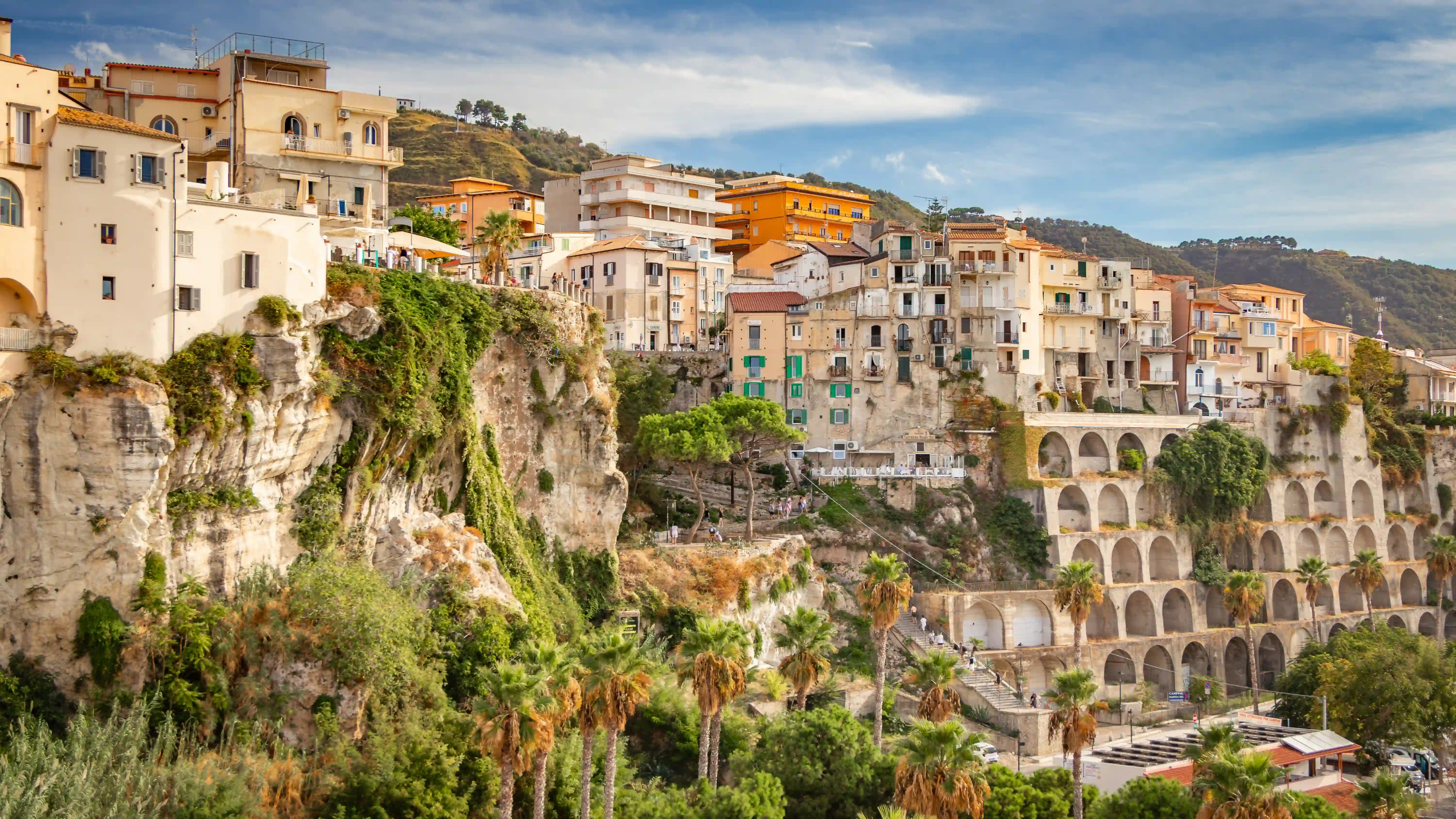 Buildings on a rocky cliff with a multi-level arched structure below and palm trees in the foreground.