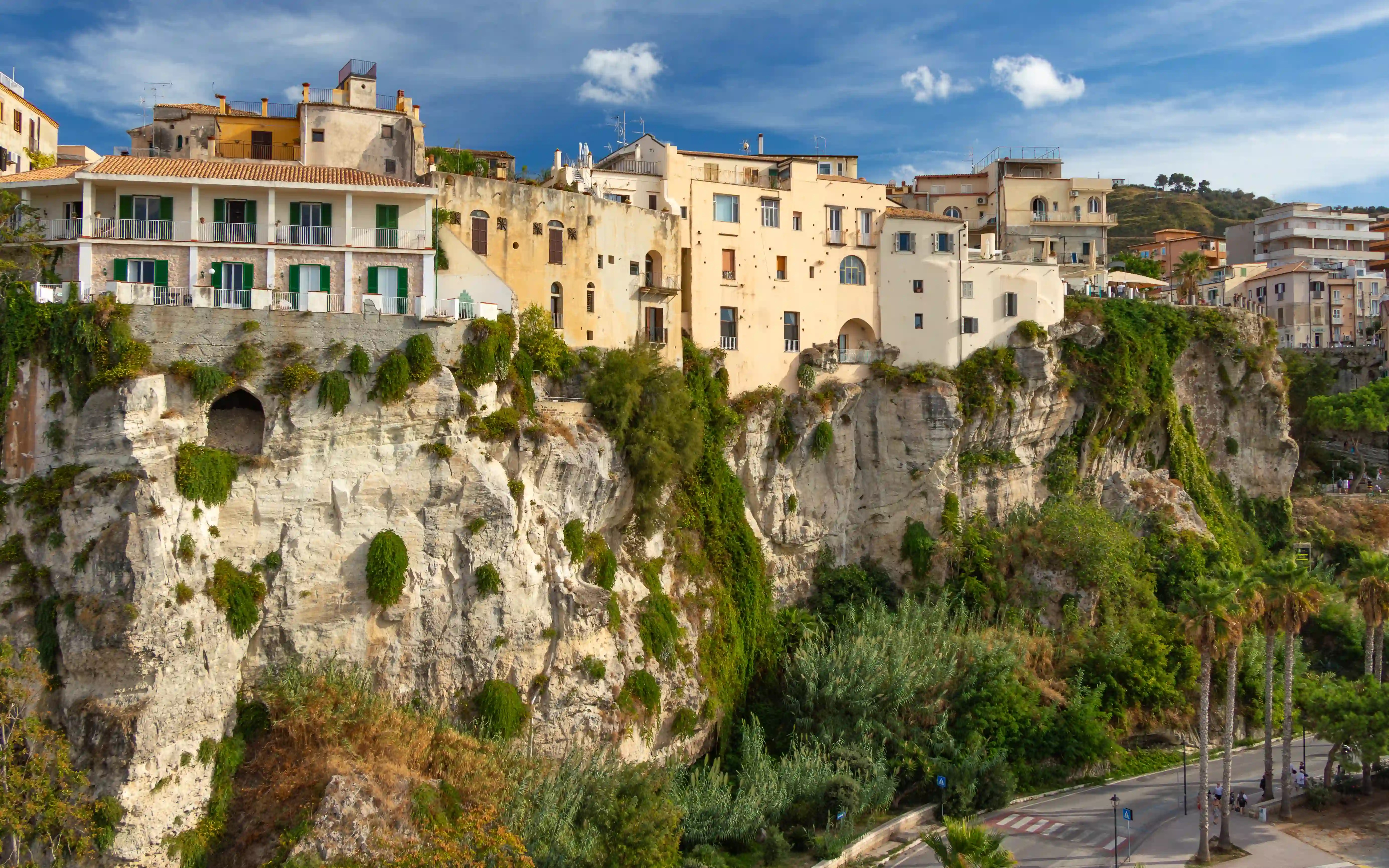 Light-colored buildings with balconies sitting along a steep rocky cliff with greenery.