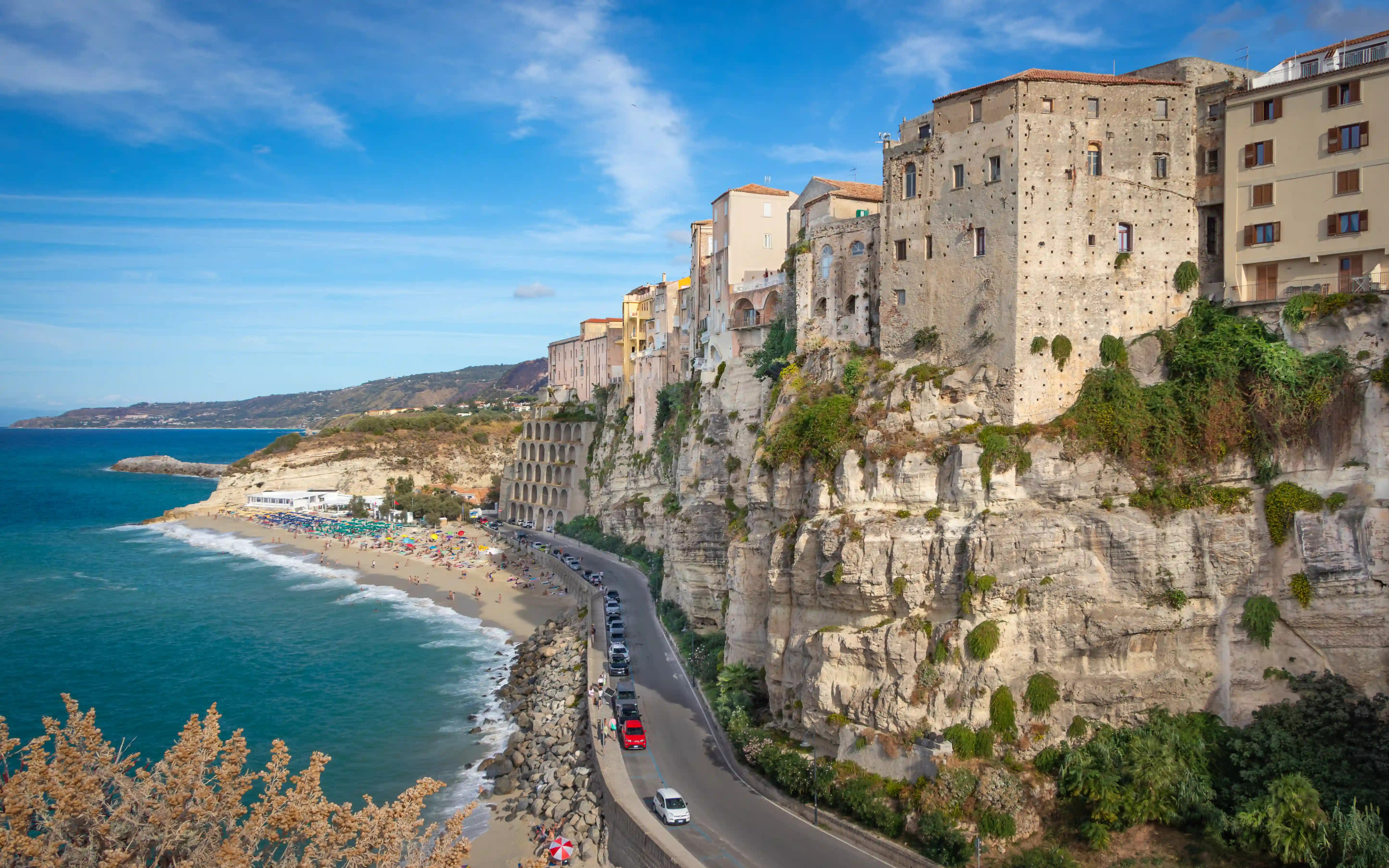Coastal road running along a beach with cliffs and buildings rising above the shoreline.