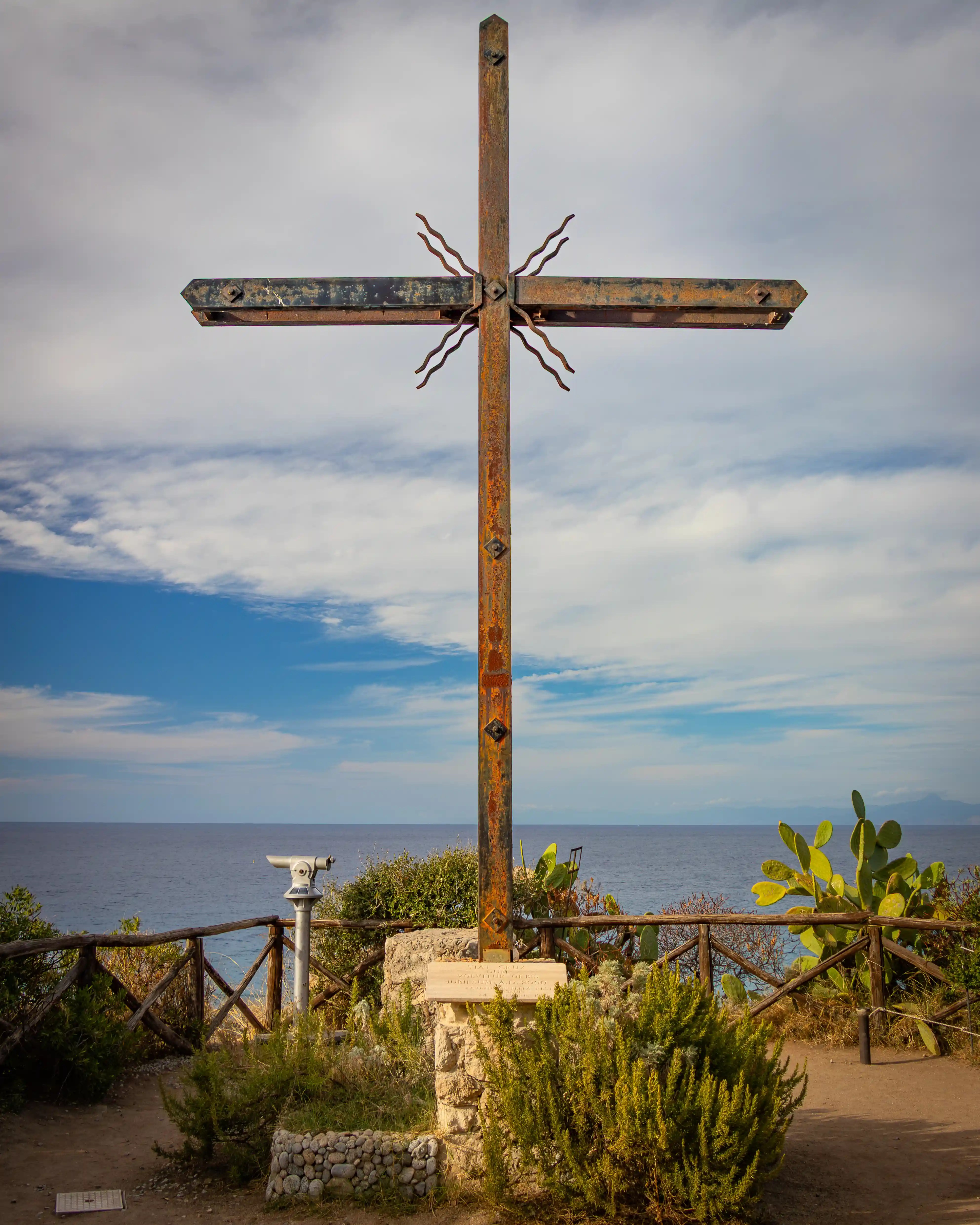 Metal cross on a stone base overlooking the sea with a viewing platform and plants nearby.