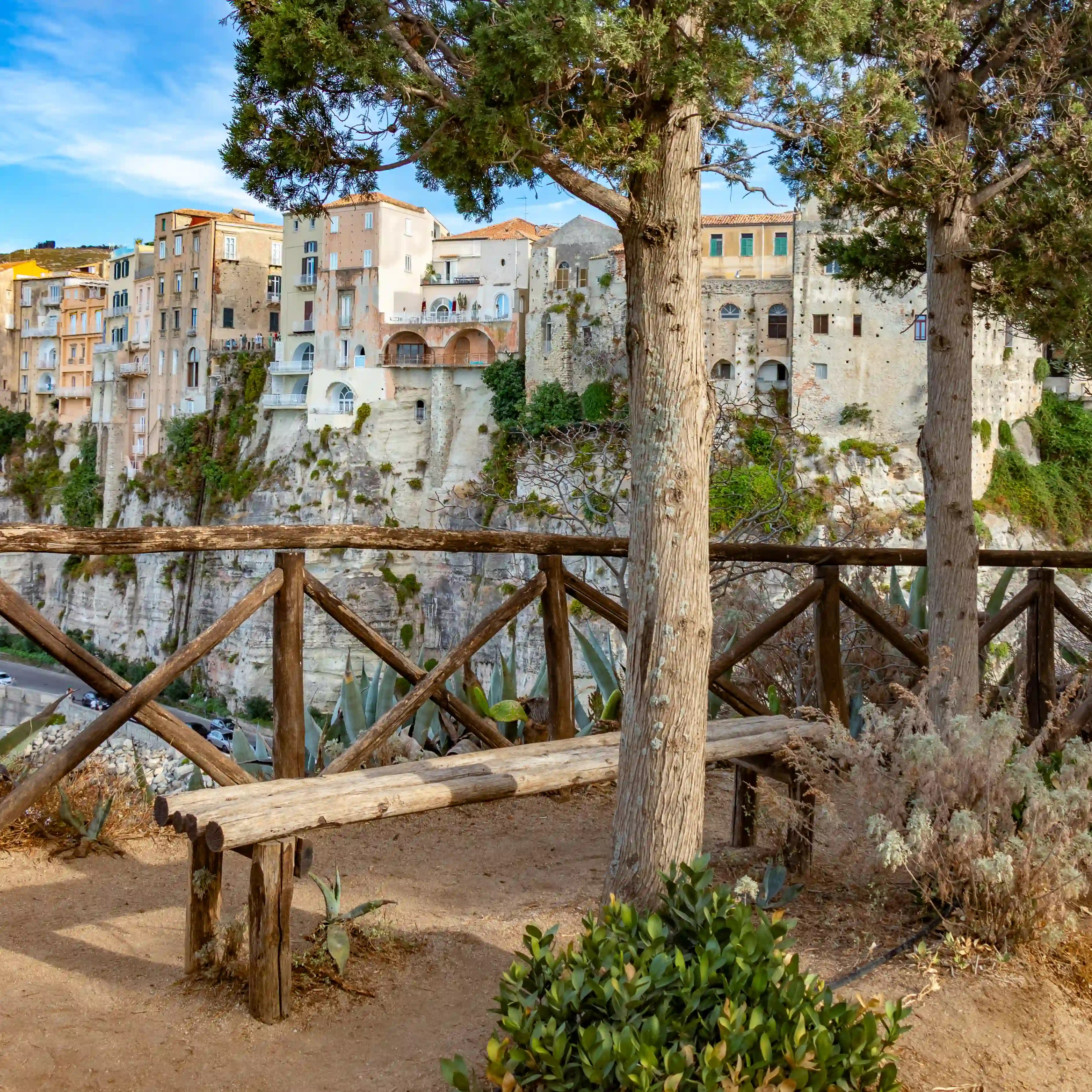 Cliffside town buildings viewed from a wooden fenced overlook with trees in the foreground.